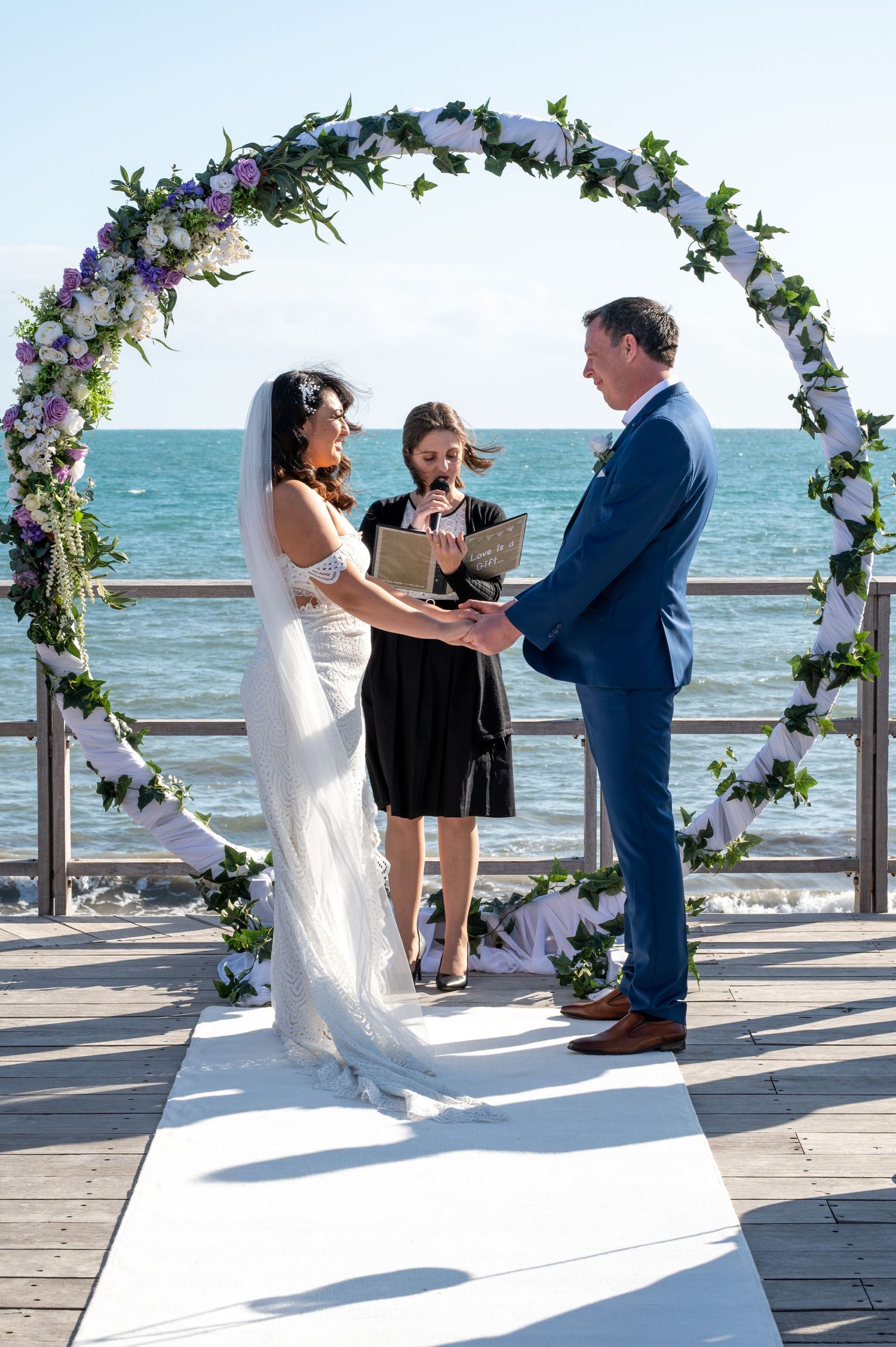 Beach wedding ceremony under white and purple floral arch with ocean backdrop at a seaside pier.
