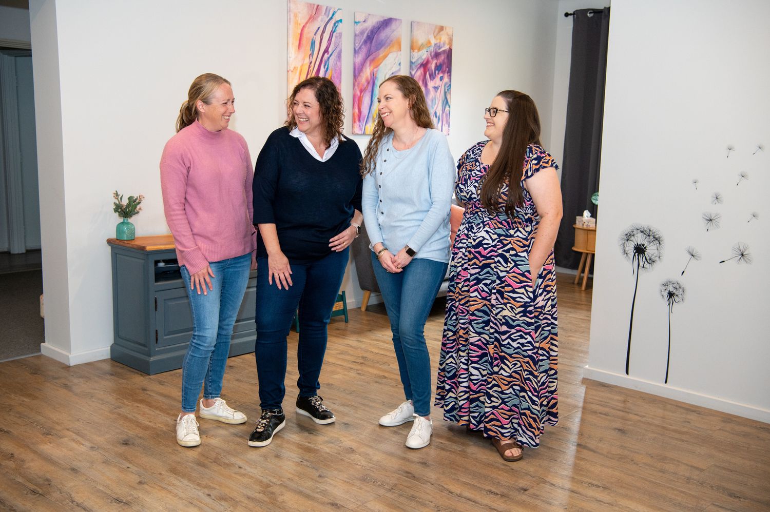 A group of people stand chatting and laughing together in a room with hardwood floors and colorful wall art.