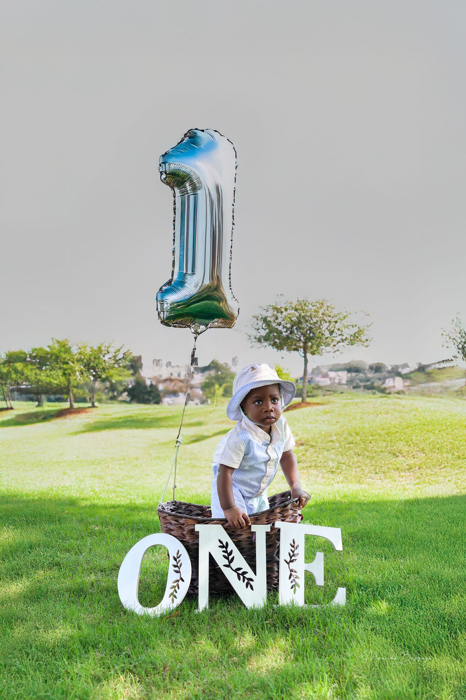 First birthday photoshoot in park with blue balloon and ONE letters on grass.