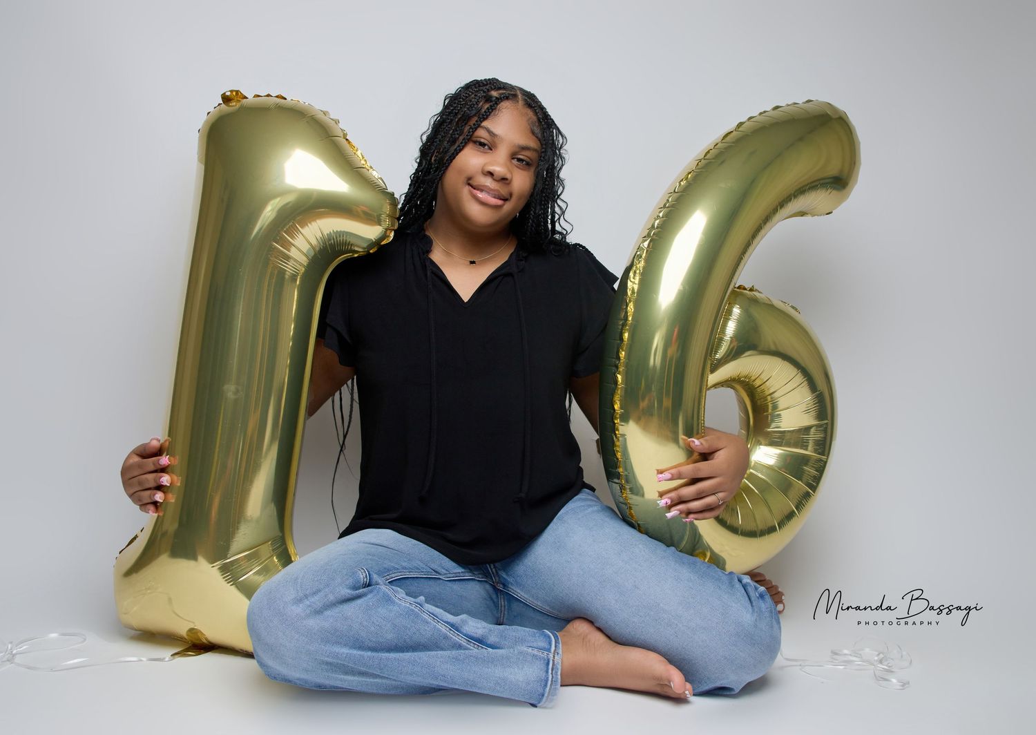 Person lounges with gold balloon numbers in casual black top and jeans.