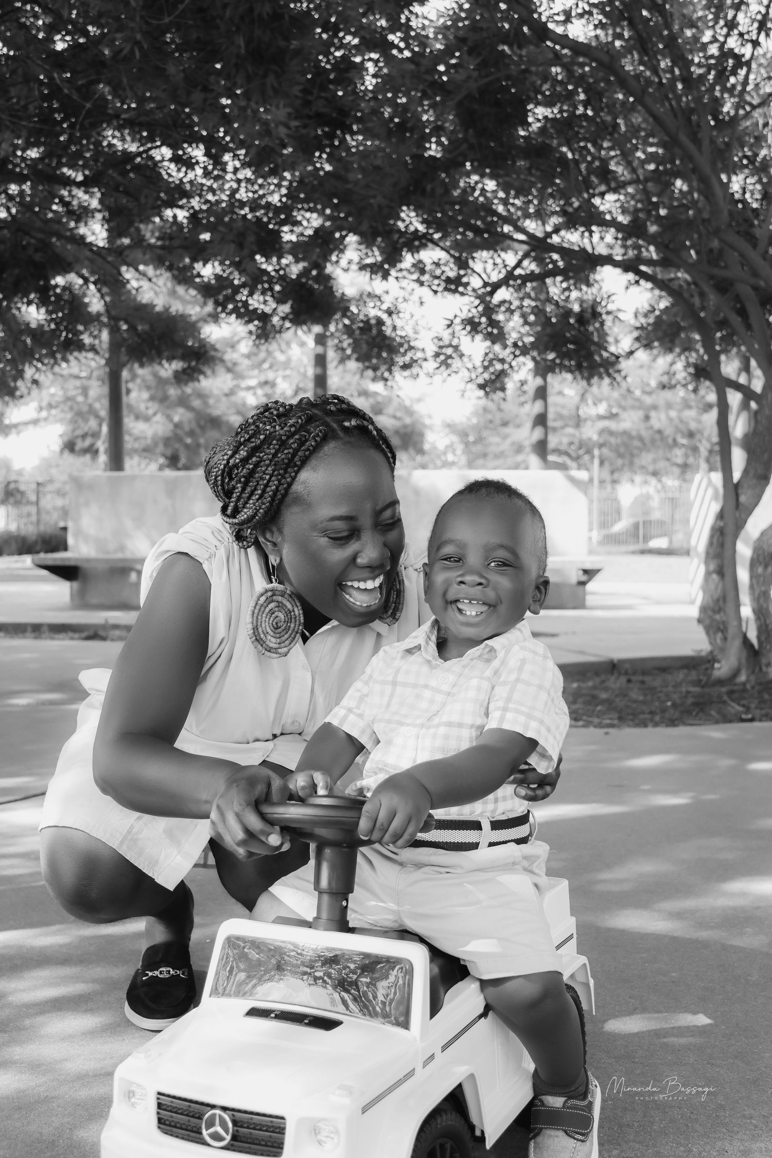 Black and white photo of joyful moment with child on toy Mercedes car in park setting.