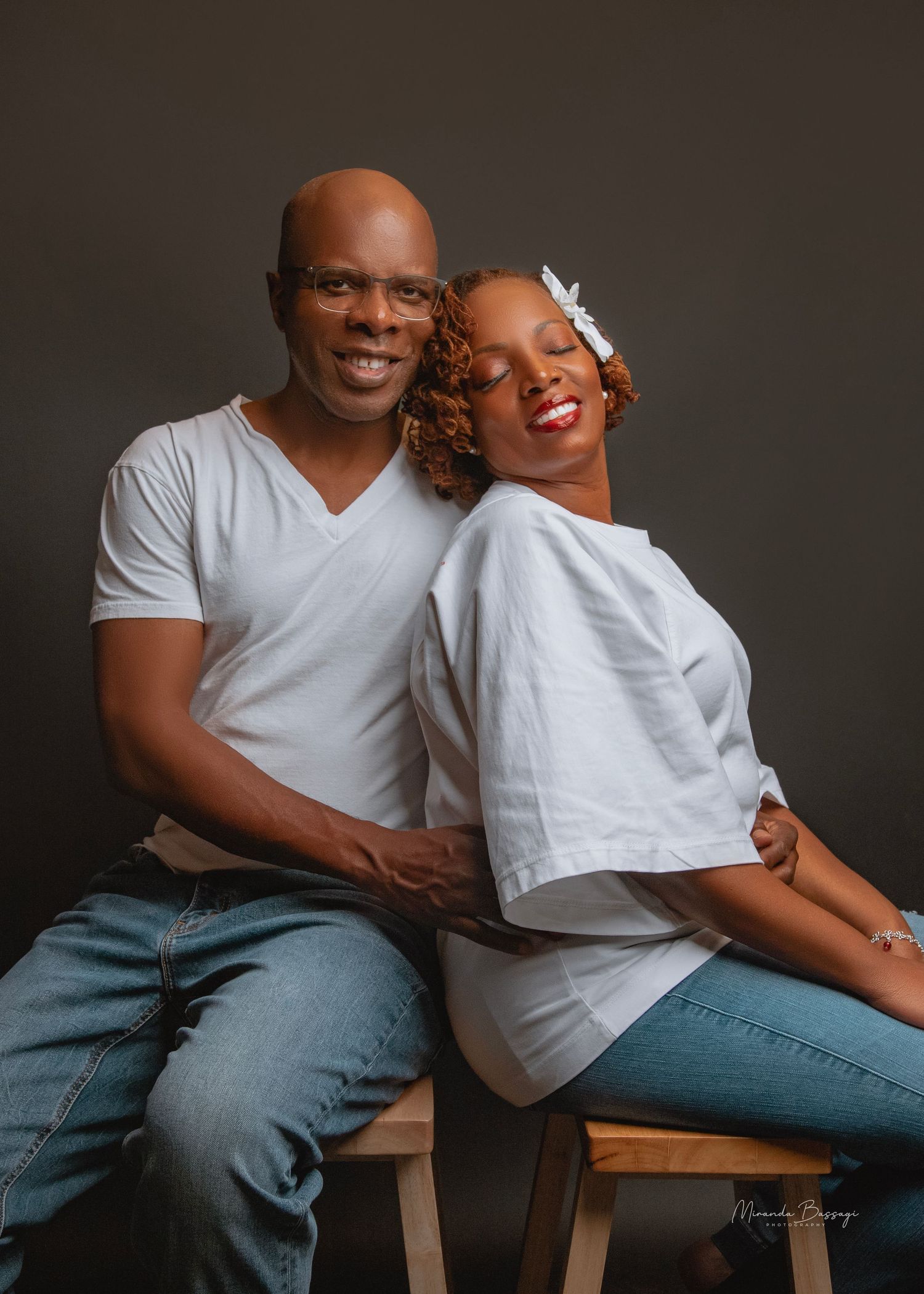 Couple in white casual wear posing together on wooden stool in studio setting.