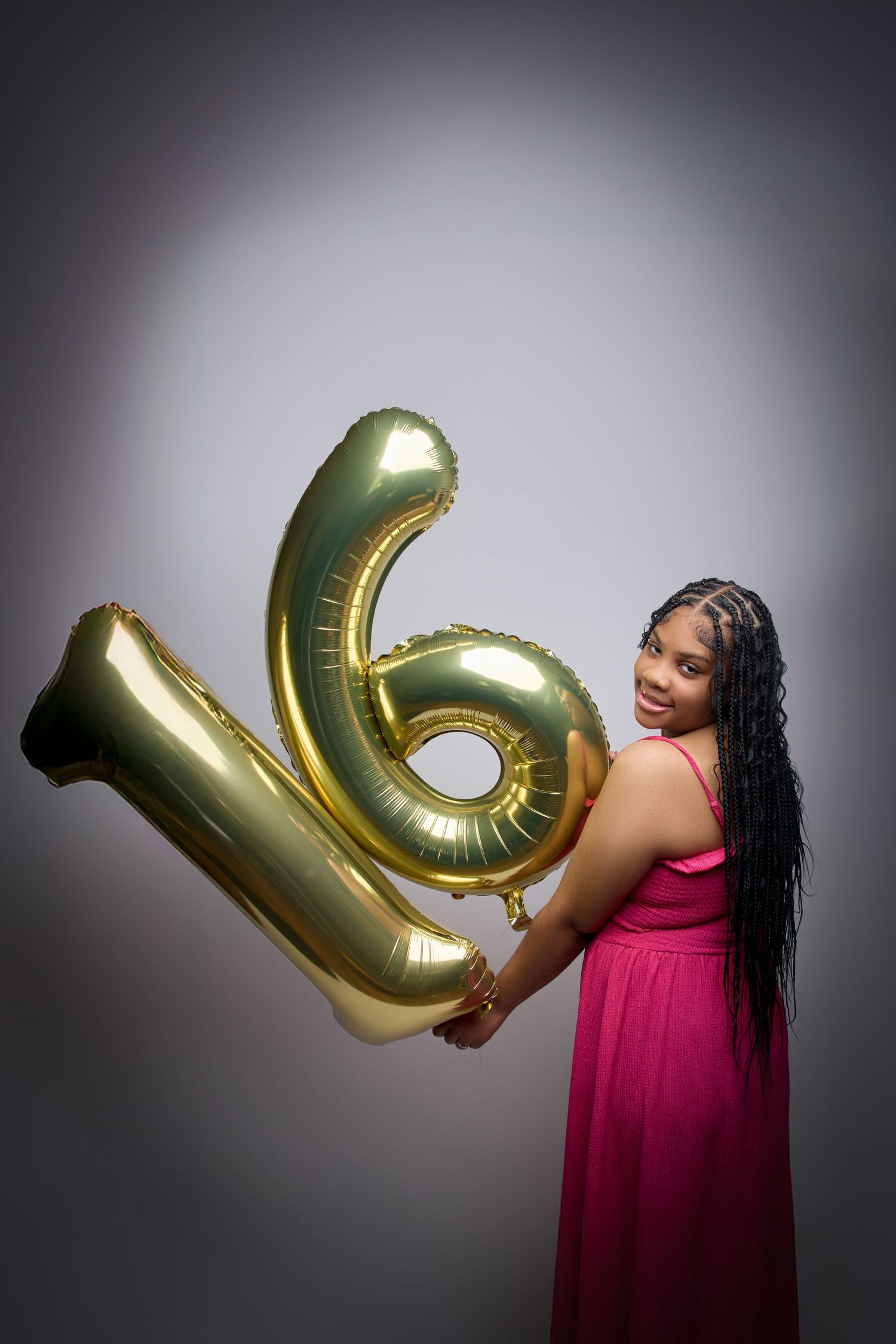 Person in pink dress poses with large gold balloon numbers against dark backdrop.