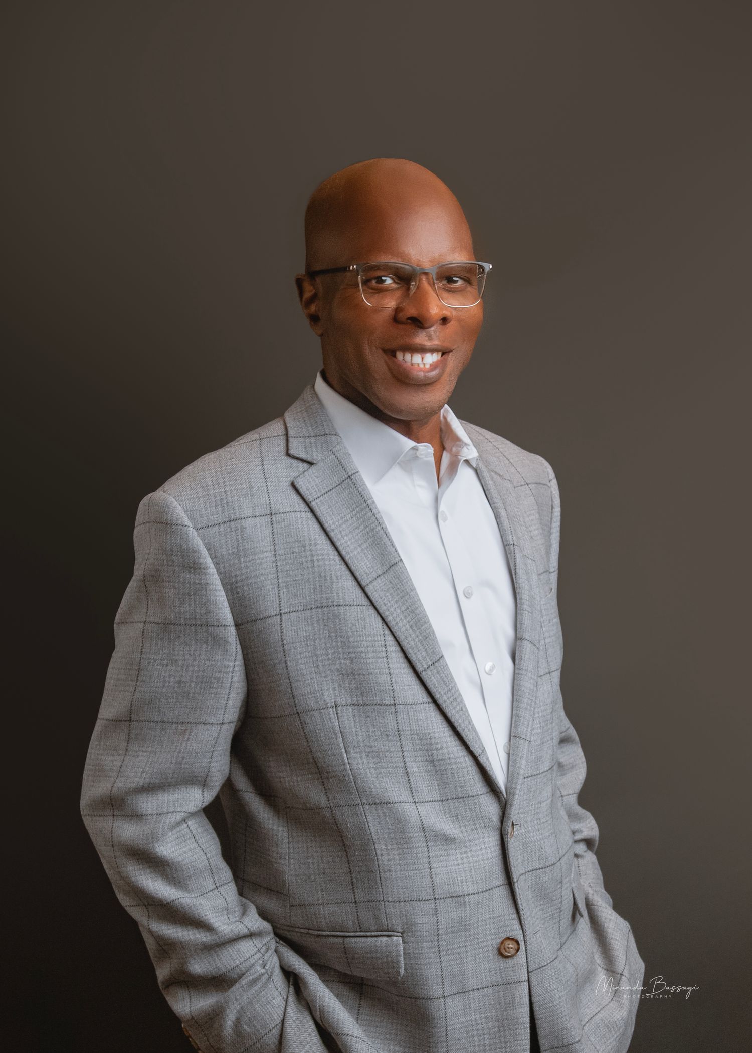 Professional headshot of person wearing grey plaid blazer and glasses against neutral background.