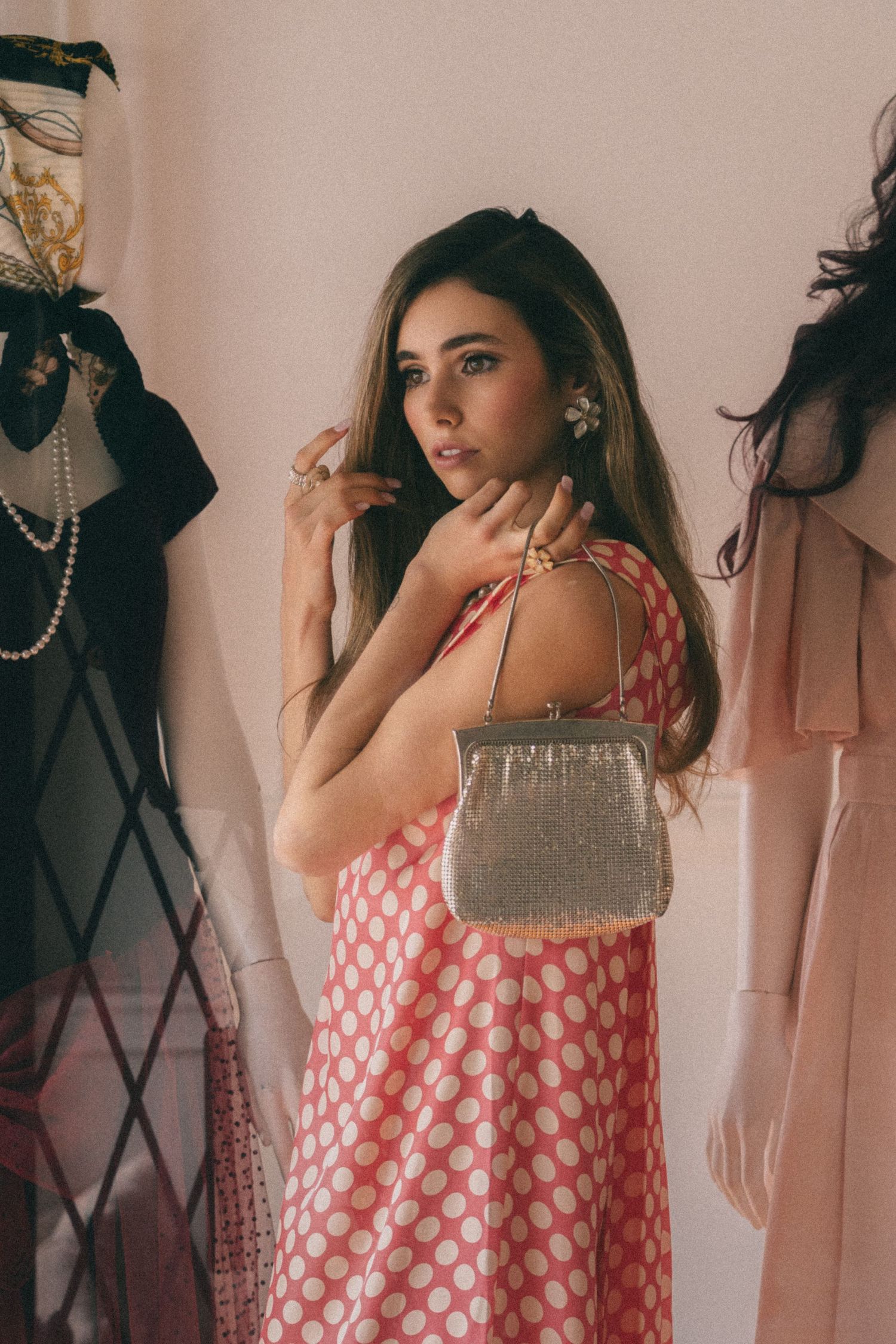 A person poses in a pink and white polka dot summer dress with a metallic handbag against a light colored wall.