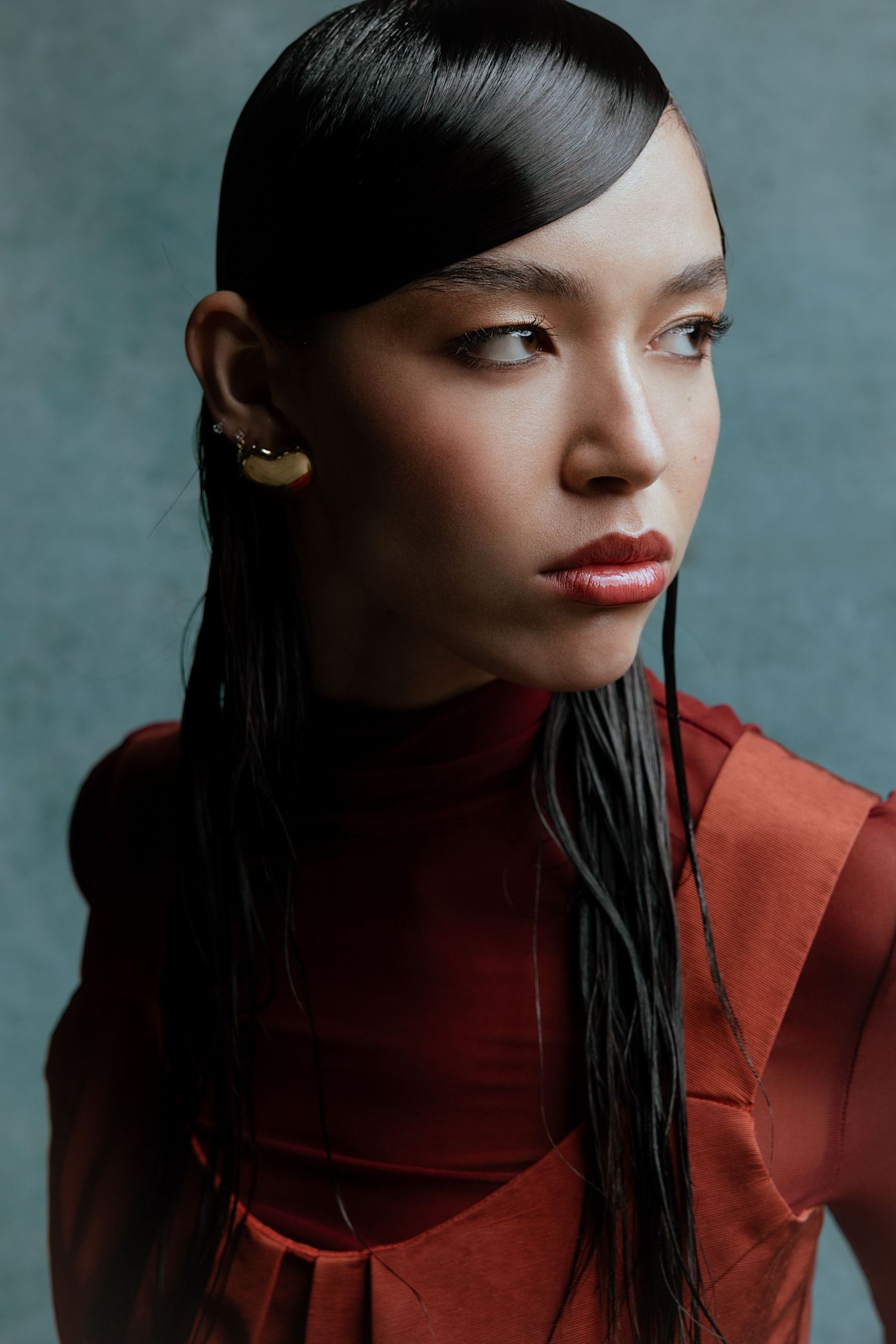 Dramatic side profile portrait with red lipstick and sleek hair against teal background.