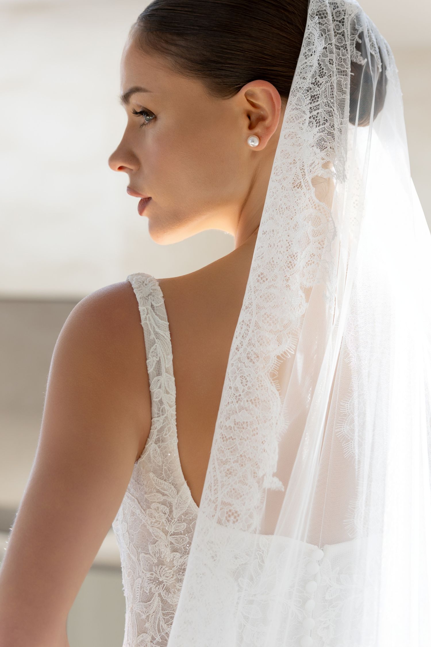 Side profile view of a bride wearing a delicate lace wedding gown and flowing veil with pearl earrings.