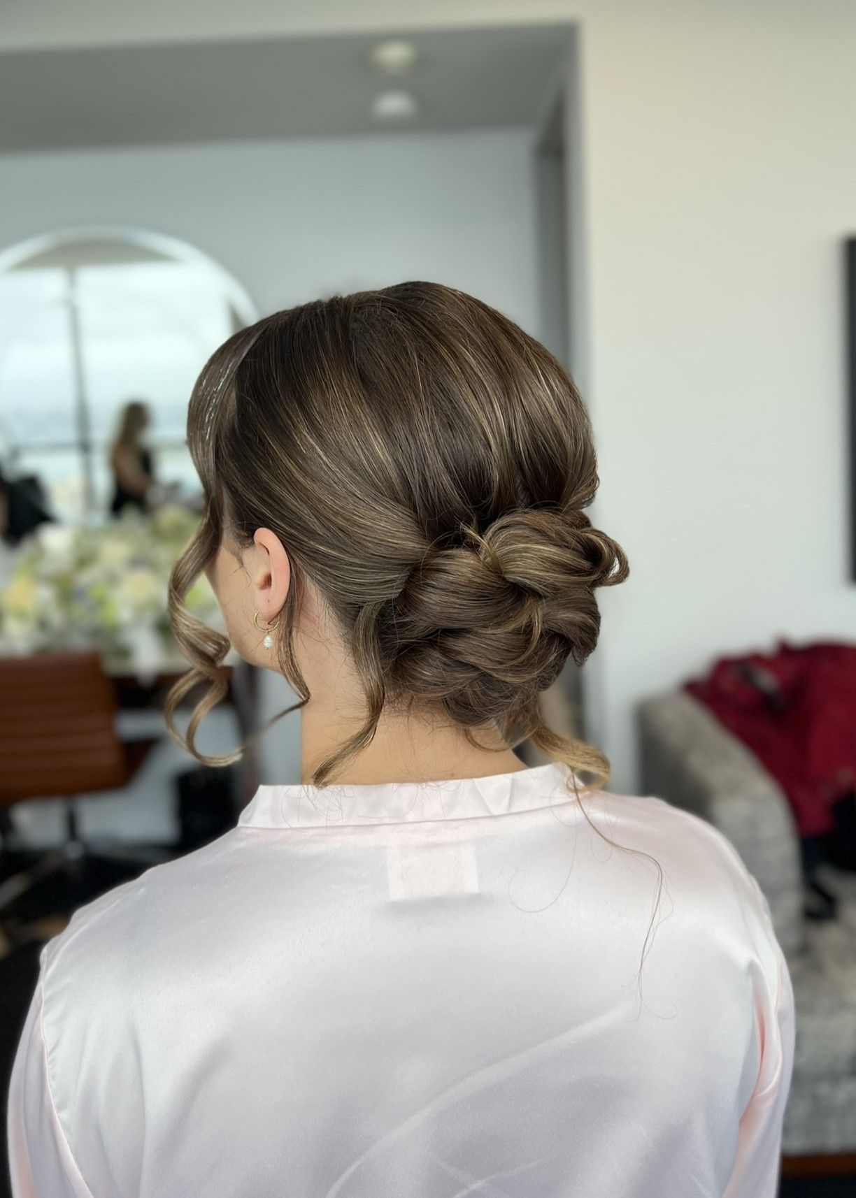 Elegant updo hairstyle with twisted bun and loose strands shown from behind on person wearing light pink silk top.