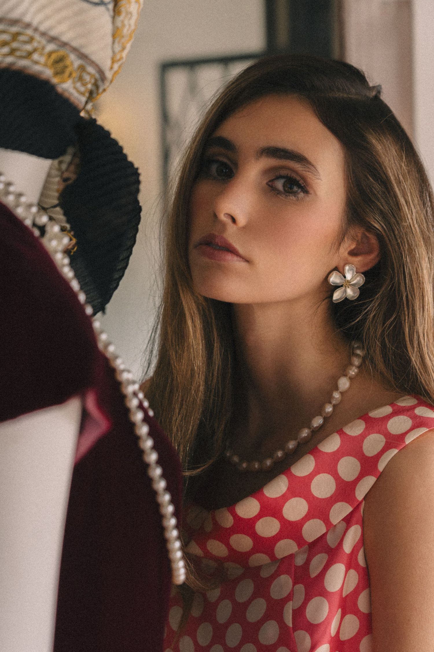 A stylish portrait featuring a pearl necklace and red polka dot dress against a dark background.