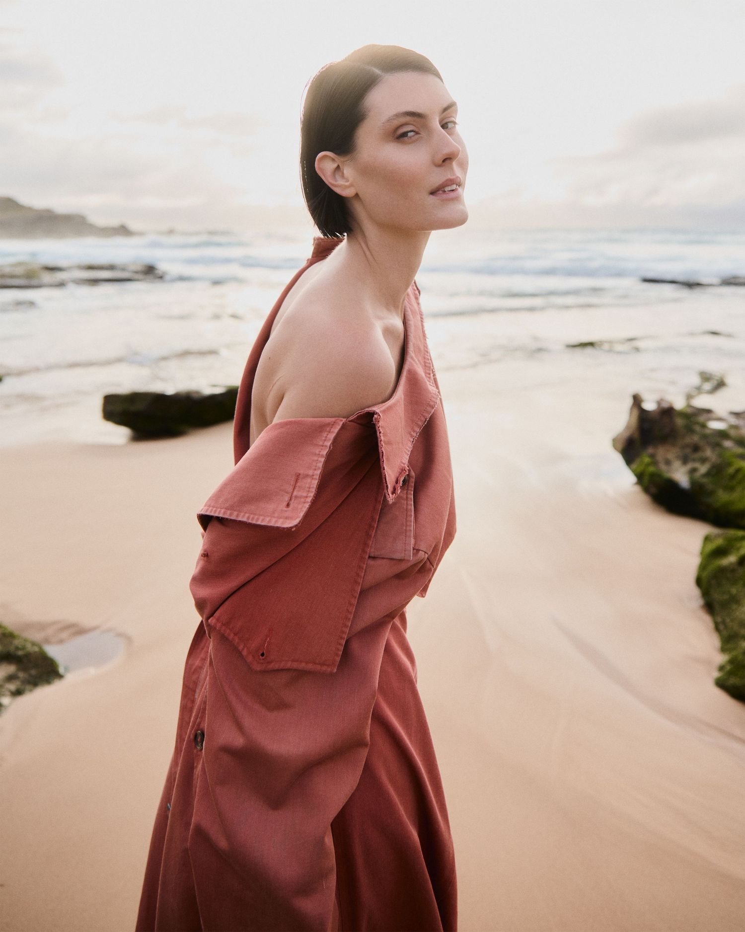 A person in a rust-colored jacket walks along a misty beach with waves and rocks in the background.