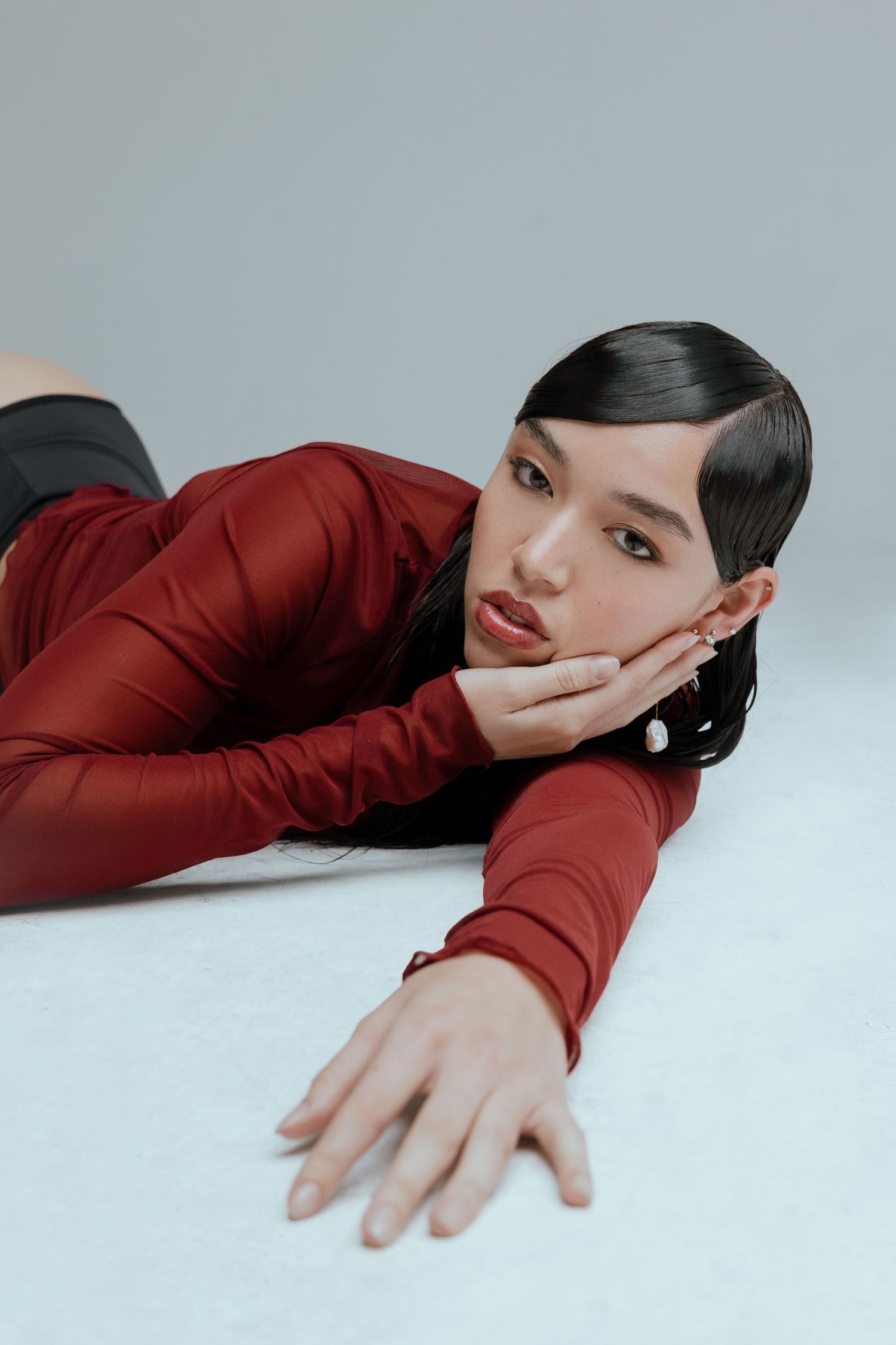 Fashion model in red long-sleeved top poses elegantly on white background with dramatic makeup and sleek hairstyle.