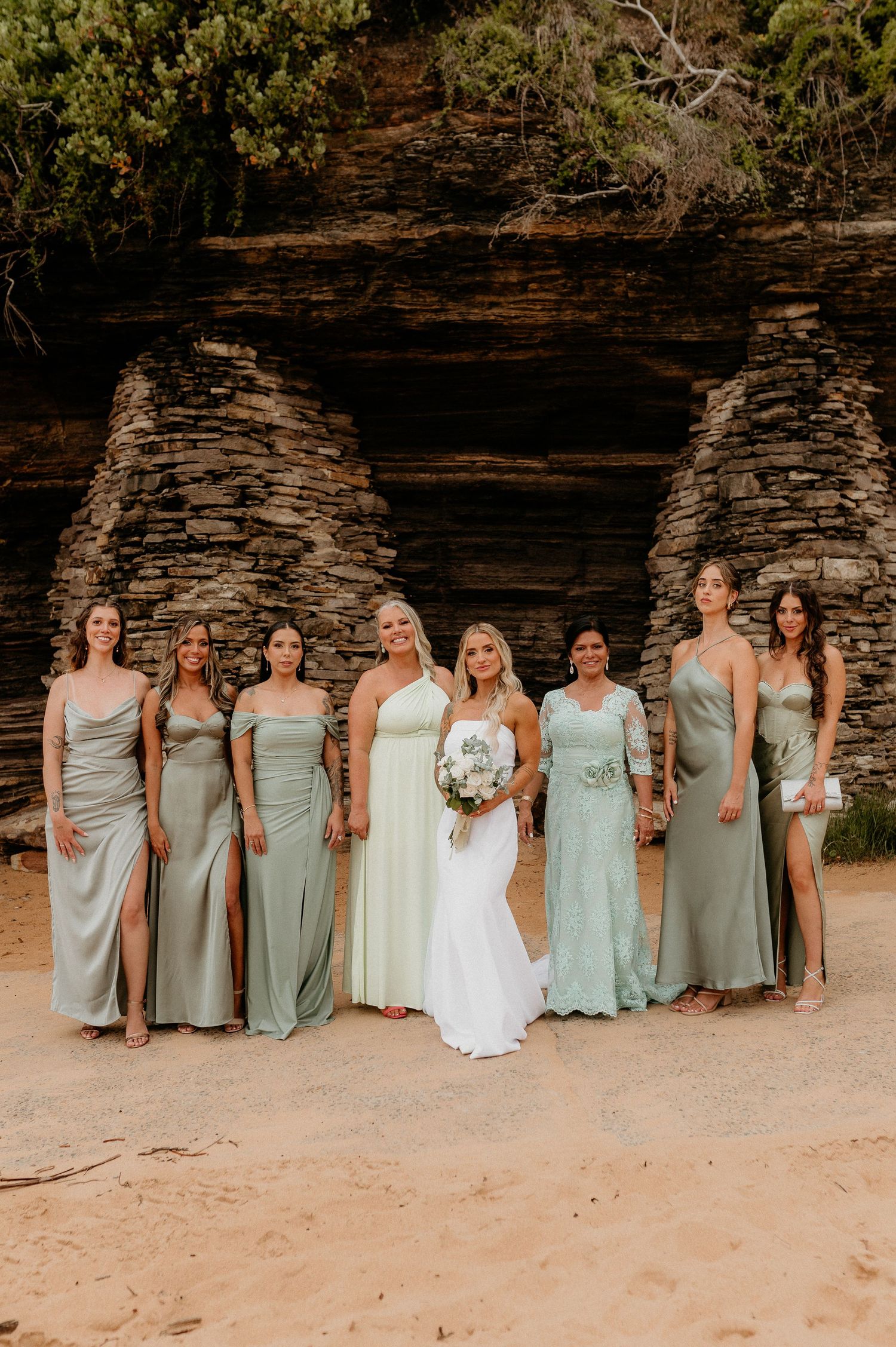 Wedding party in sage green bridesmaid dresses posed on beach with stone wall backdrop.