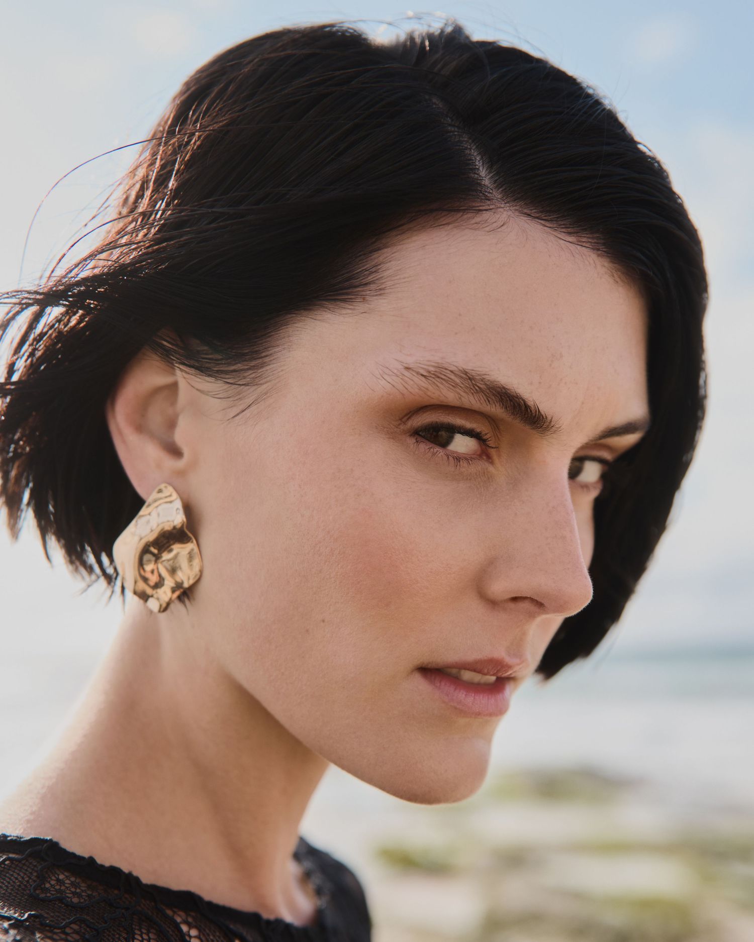 Portrait of woman with dark pixie haircut wearing gold earrings against blurred beach background.