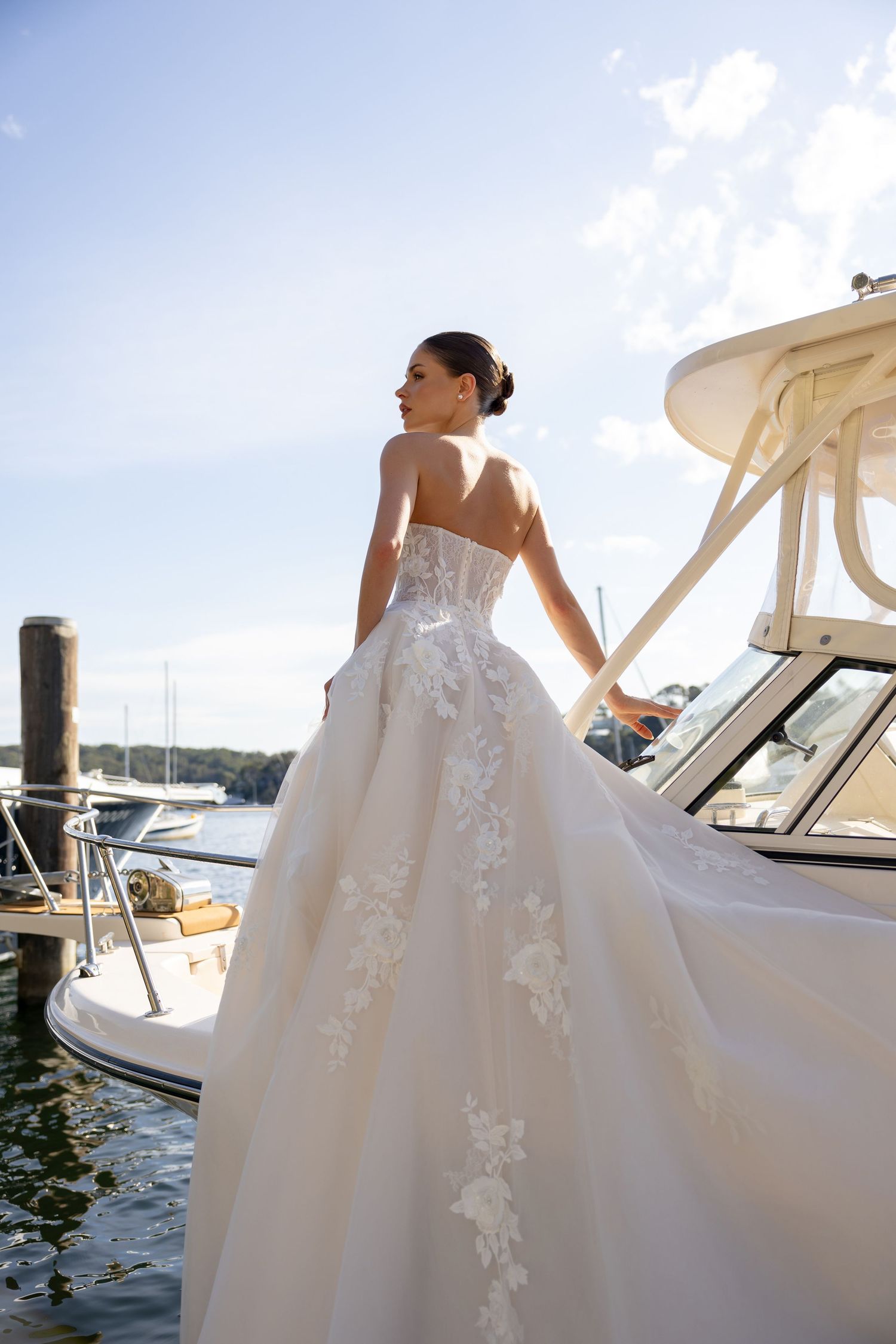 A bride in a flowing white ball gown with floral appliques poses on a dock with boats in the marina background.
