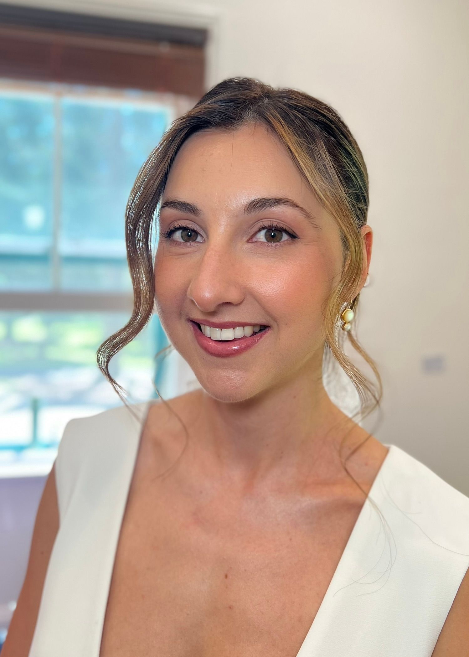A smiling person wearing a white sleeveless top and gold earrings poses for a portrait indoors.