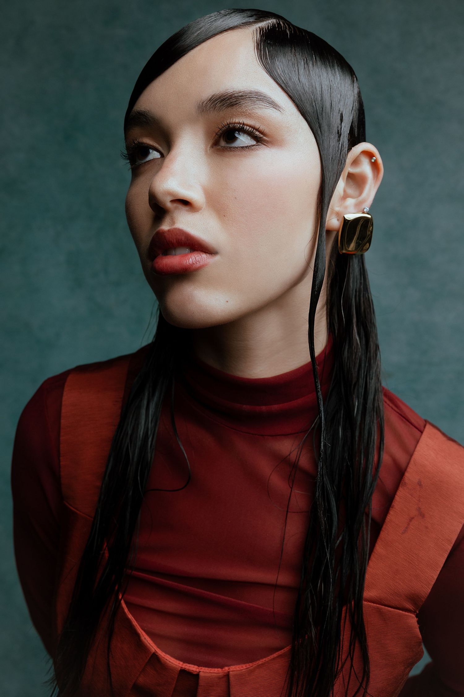Editorial portrait featuring red turtleneck top and sleek dark hair against moody green backdrop.