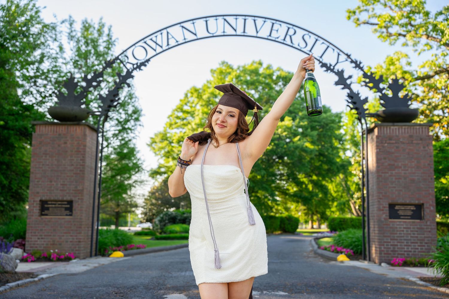 Female graduate in white dress celebrating at Rowan University entrance gate.