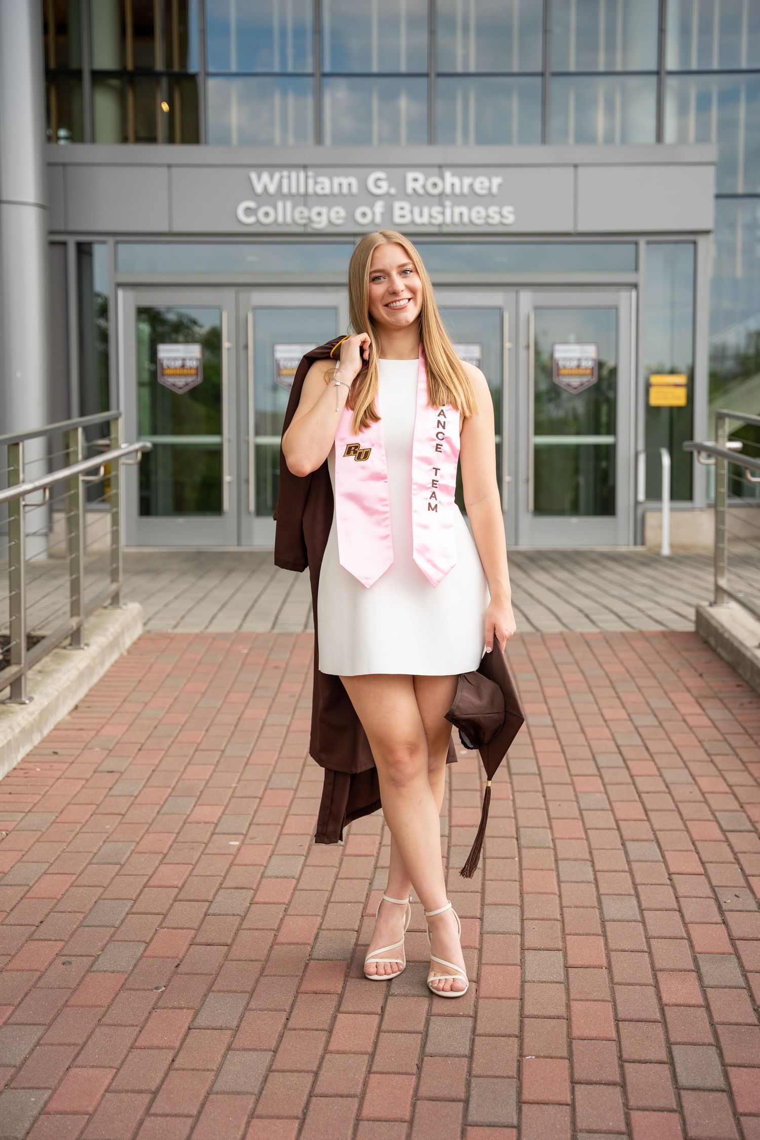 Smiling graduate in white dress with pink sash outside William G. Rohrer College of Business.