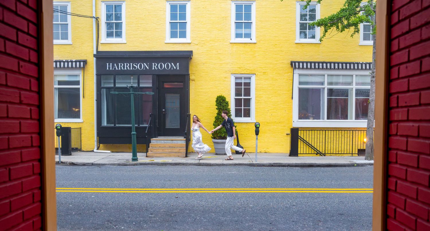 Street scene viewed through brick arch showing yellow Harrison Room building facade.