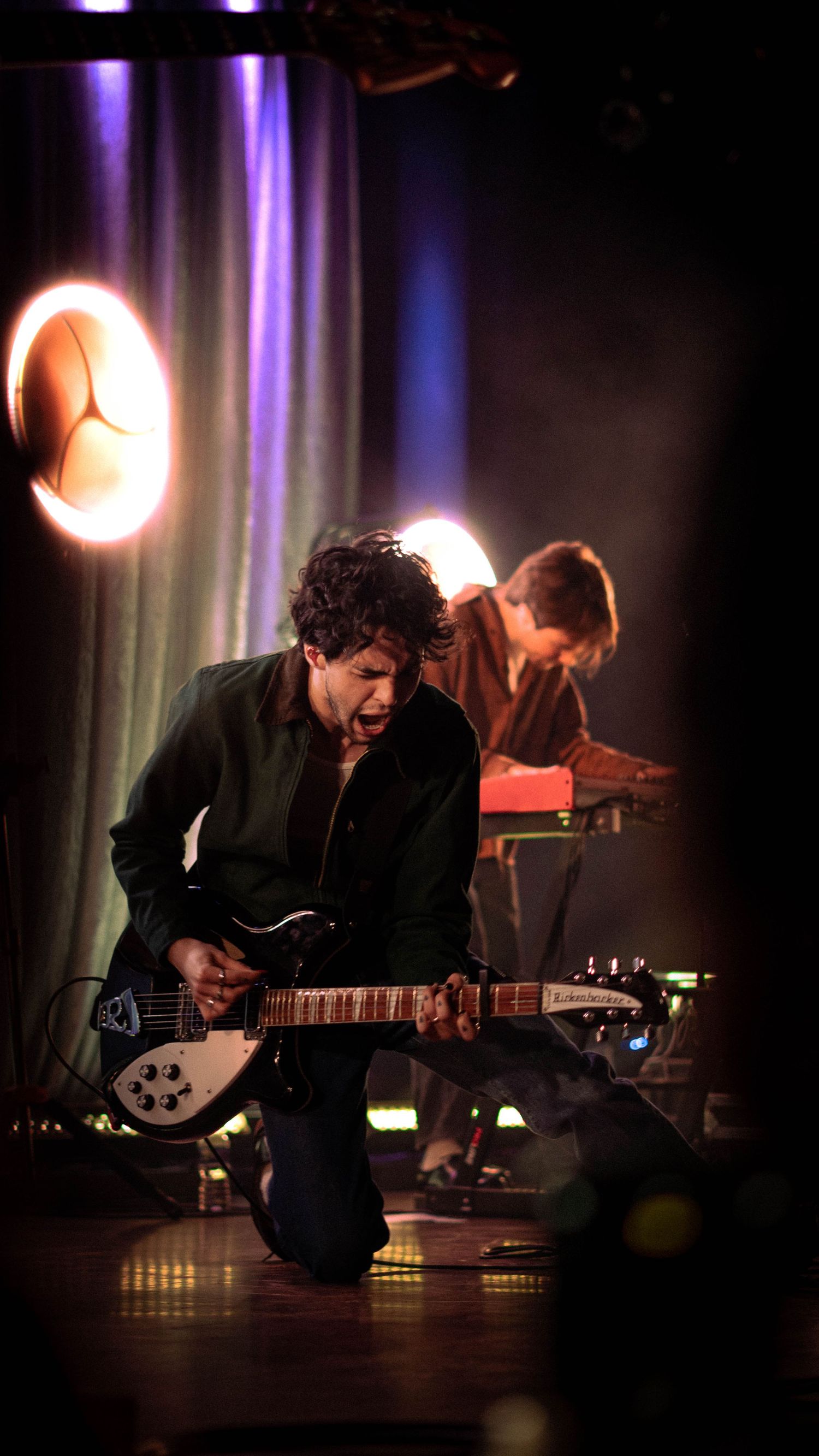 Male guitarist kneeling on stage playing electric guitar passionately with a keyboardist in the background.
