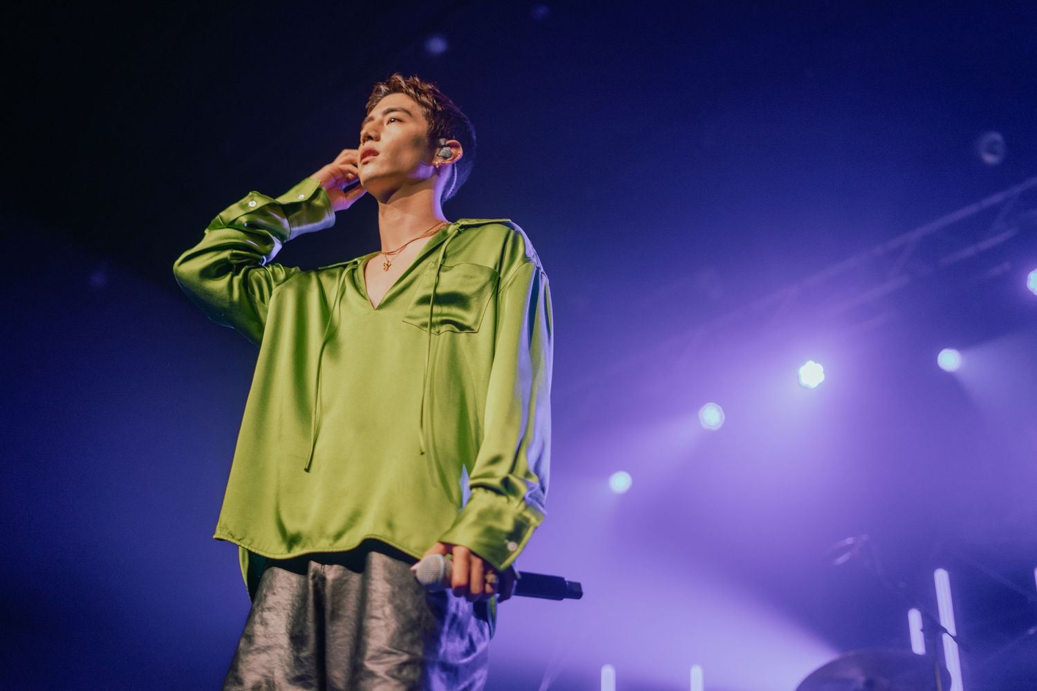 Male singer performing on stage in a green satin shirt under purple concert lighting holding a microphone.