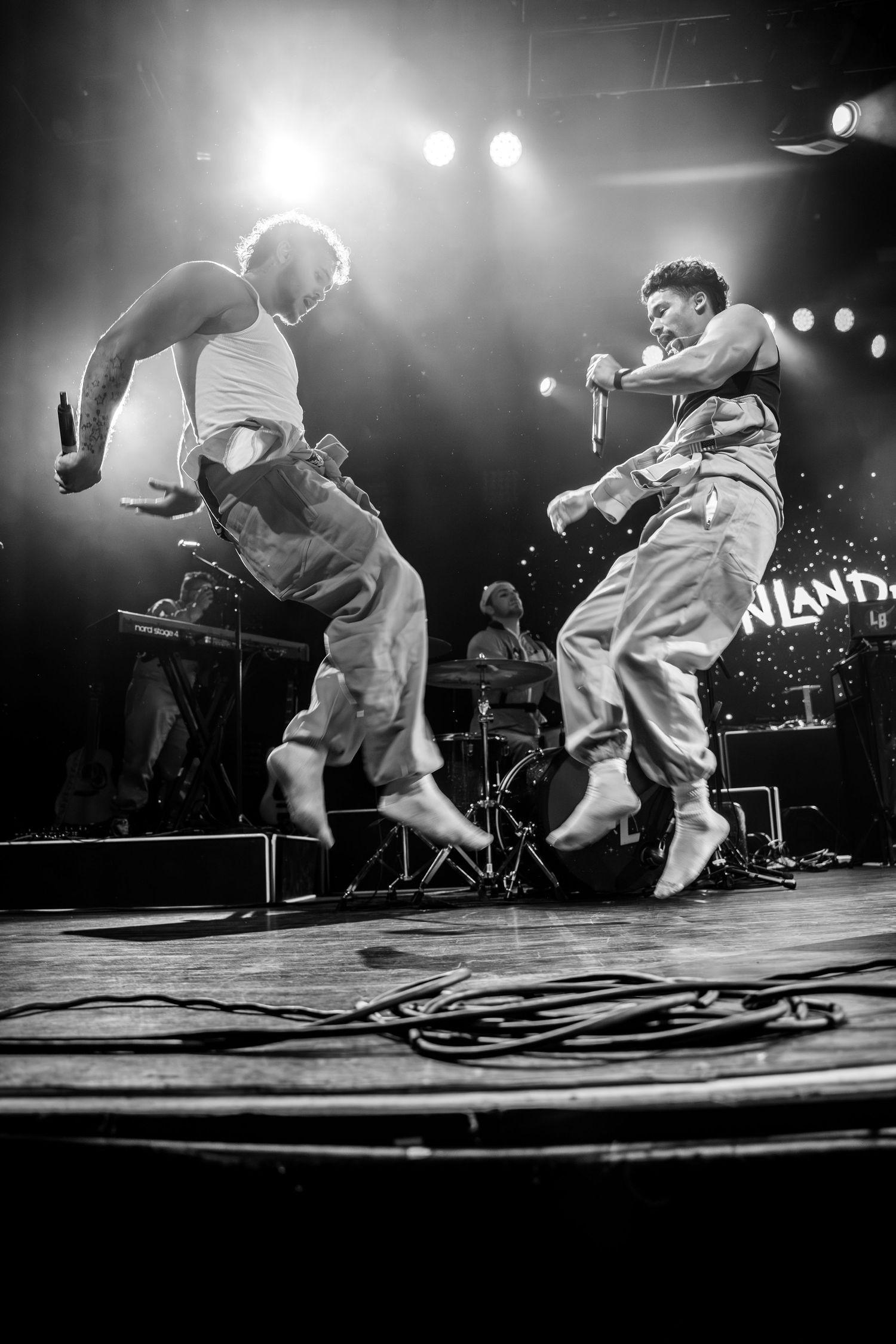 Black and white photo of two performers jumping on stage during energetic concert performance.