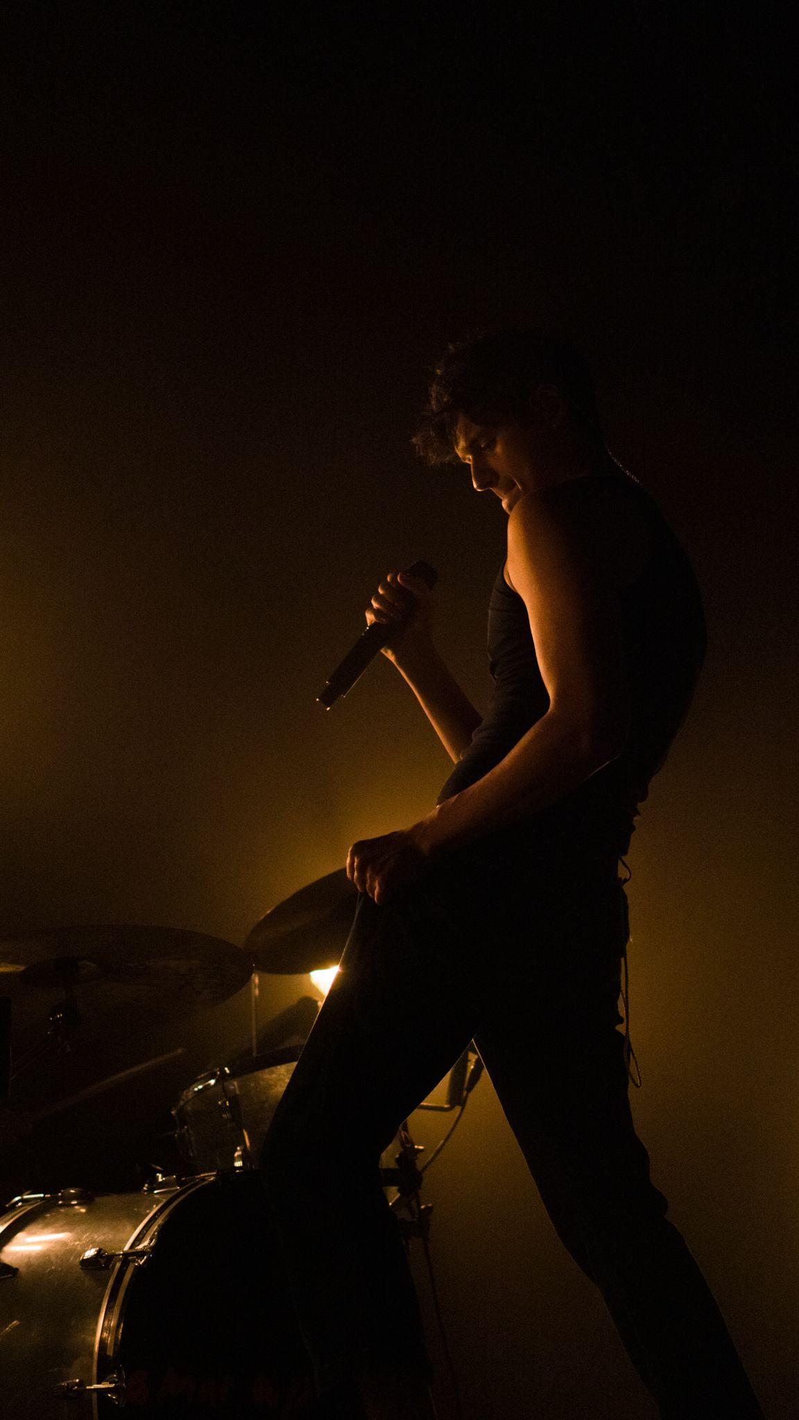 Silhouetted musician holding a microphone near a drum kit bathed in golden amber stage lighting.