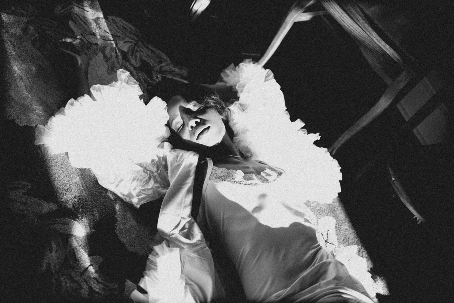 Black and white artistic photo of a bride in a white ruffled dress lying dramatically with light flares.