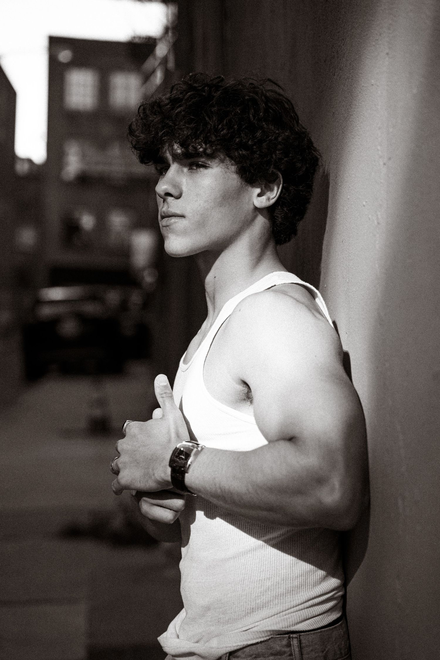 Young man in white tank top leaning against wall in black and white urban alley portrait.