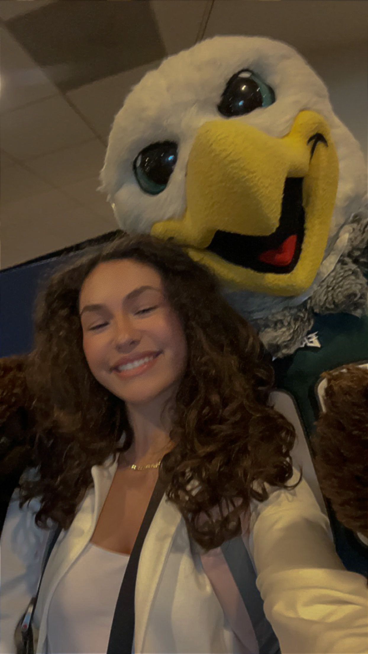 Young woman smiling in a selfie with a large eagle mascot costume at an indoor venue.
