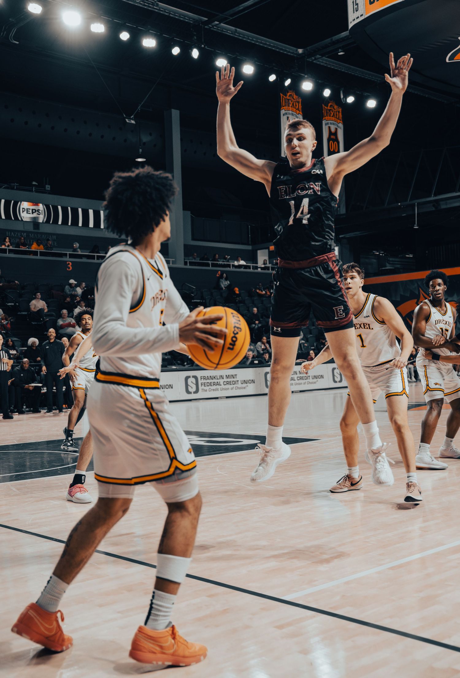 Basketball player number 14 jumps with arms raised to defend against opponent dribbling during college game.