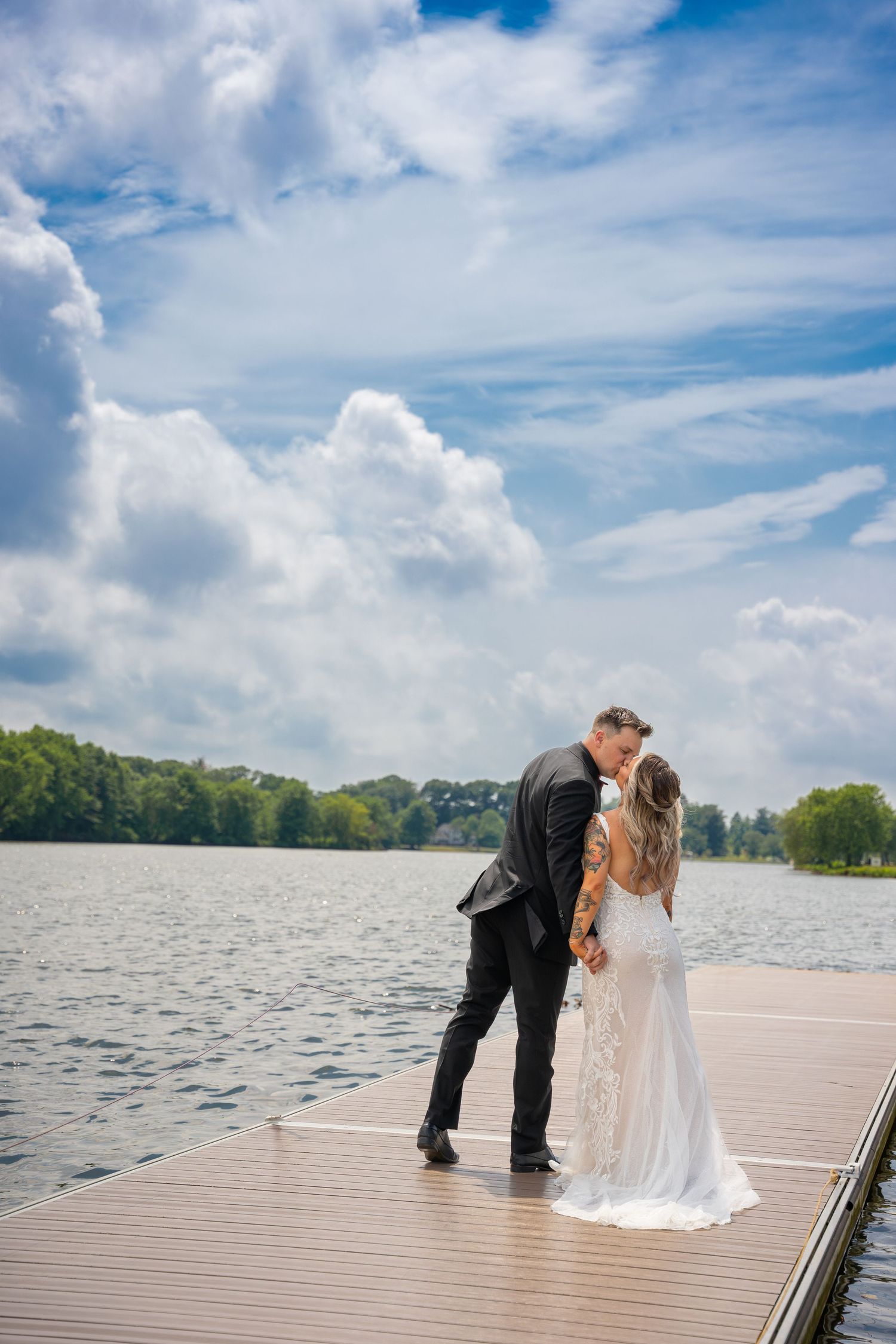 Bride and groom kissing on a lakeside dock under a cloudy blue sky.
