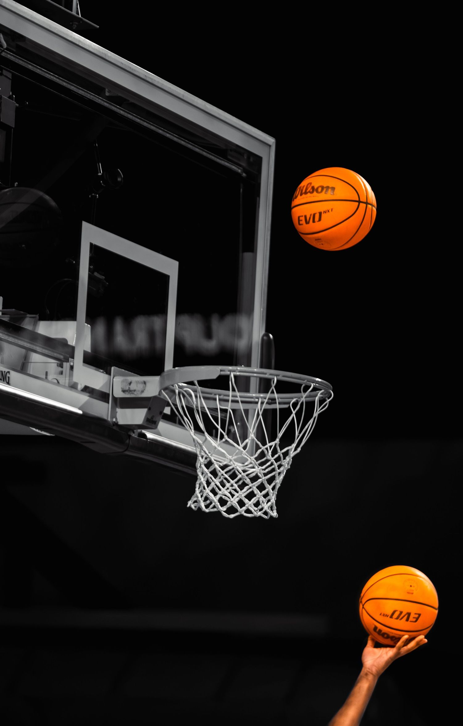 Two orange Wilson Evo basketballs near hoop and backboard against dramatic black background.