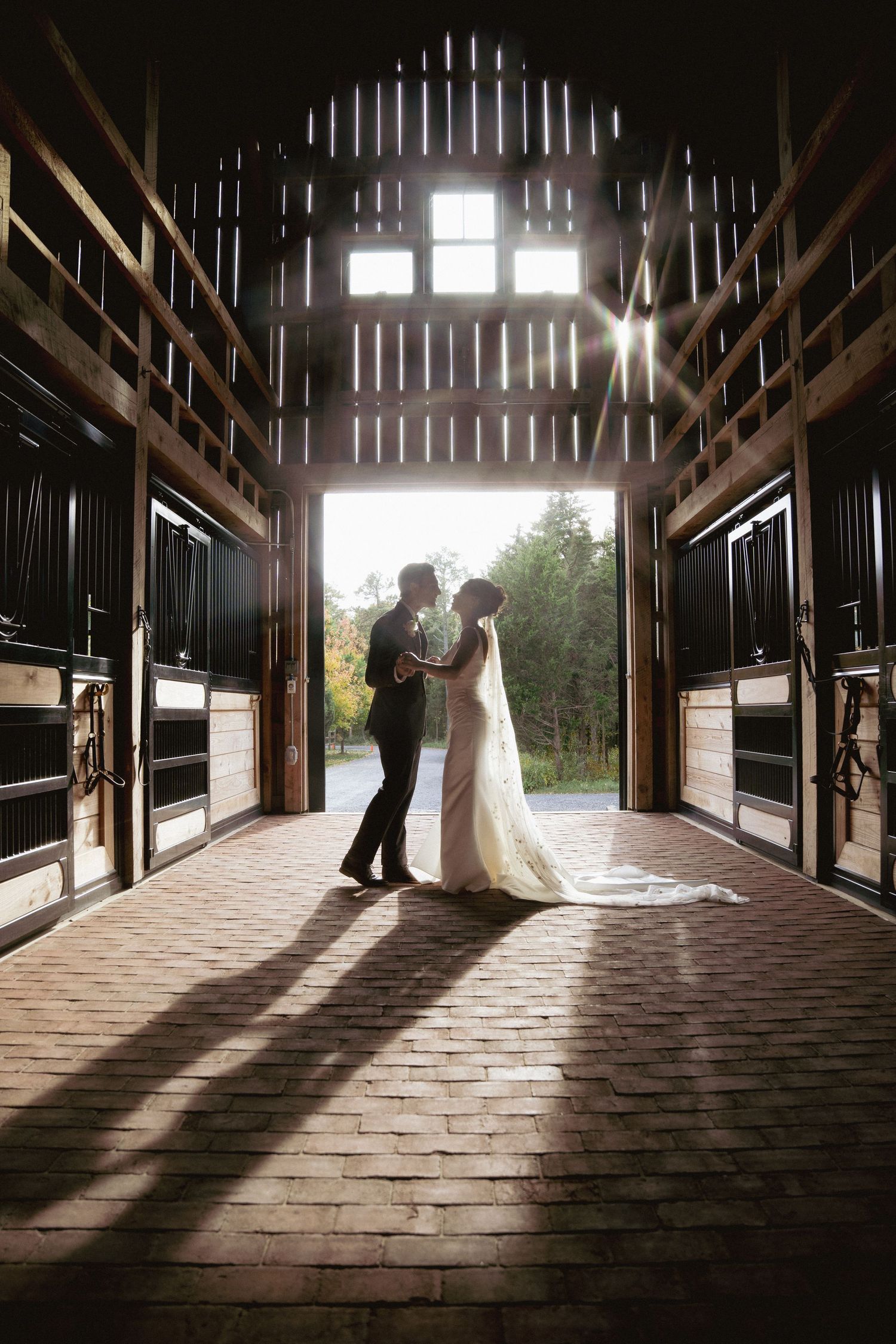 A bride and groom share a romantic moment silhouetted in the sunlit entrance of a rustic horse barn.