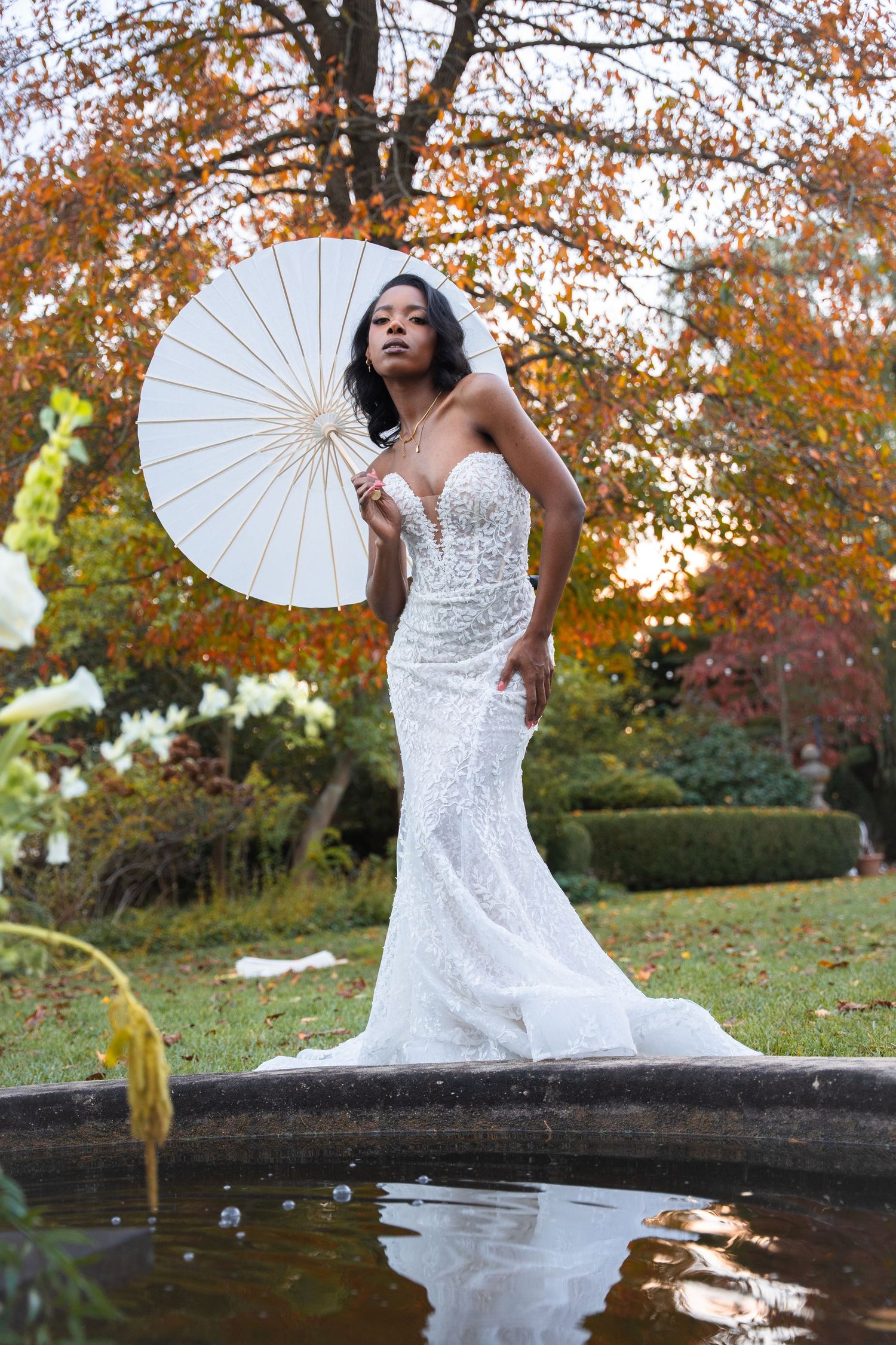 An elegant bride in a fitted beaded gown holds a white parasol by a garden fountain amid fall foliage.