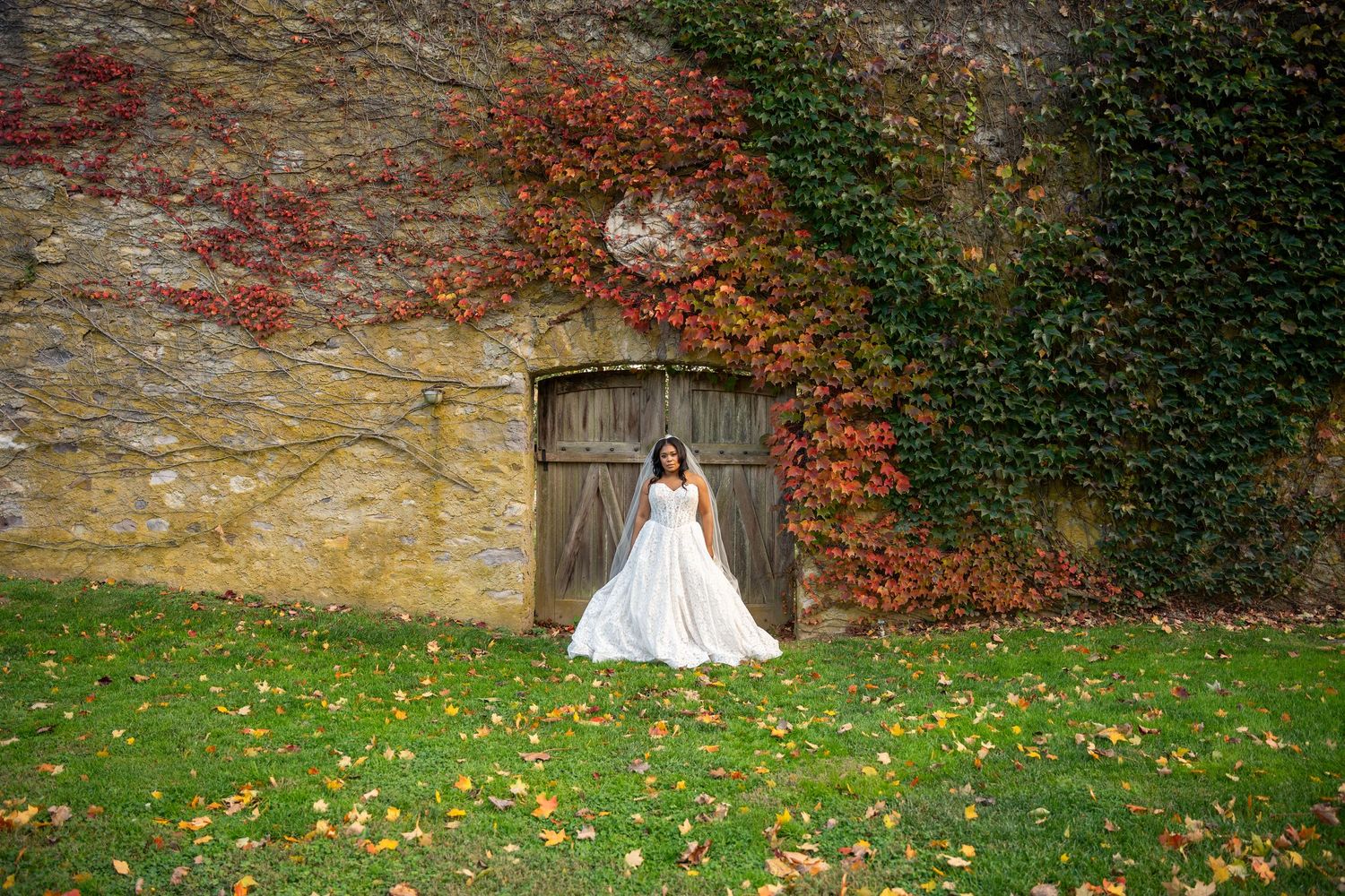Bride in white gown stands before rustic wooden doors framed by colorful autumn ivy on stone wall.