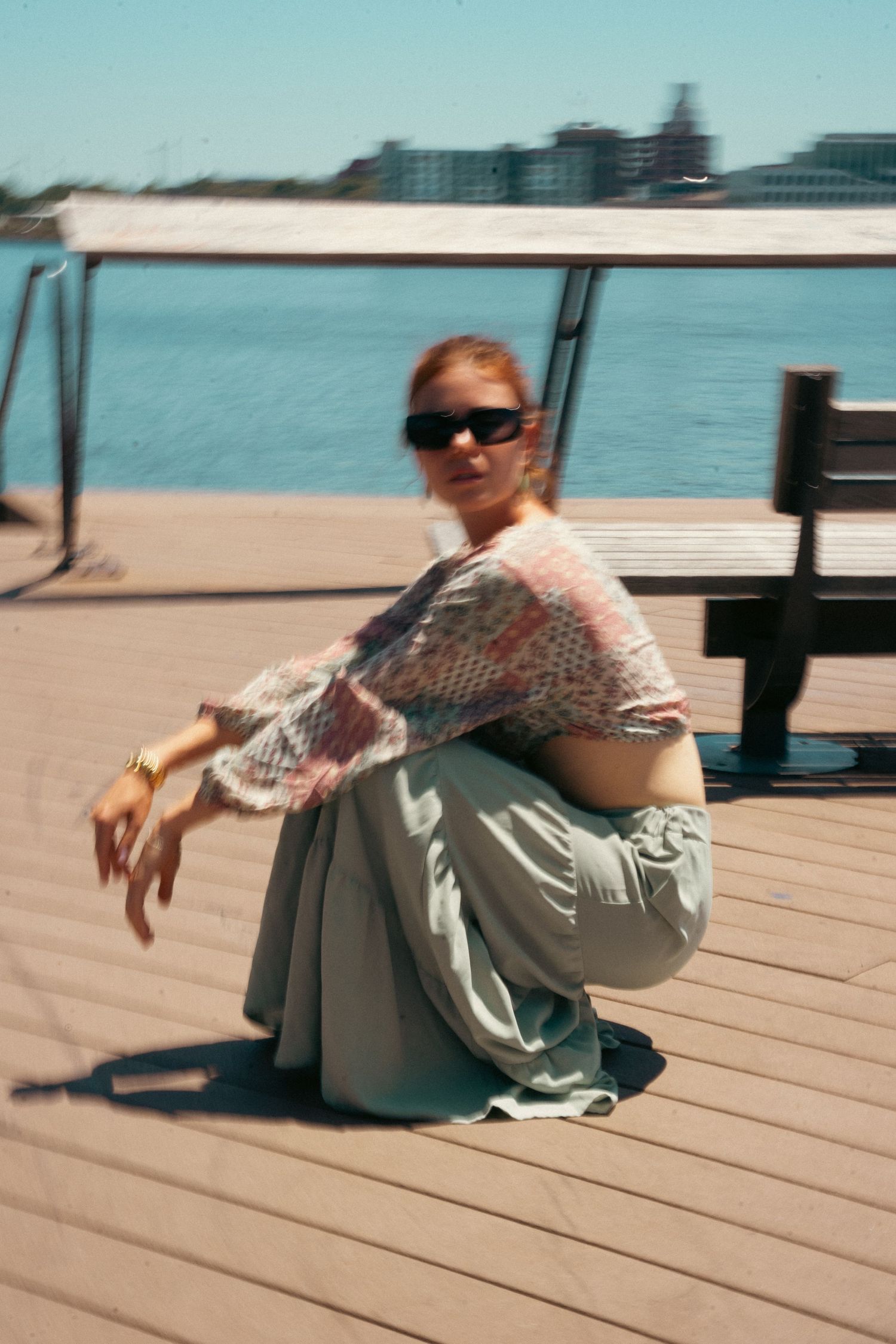 Stylish woman in sunglasses crouching on sunny waterfront boardwalk near bench.