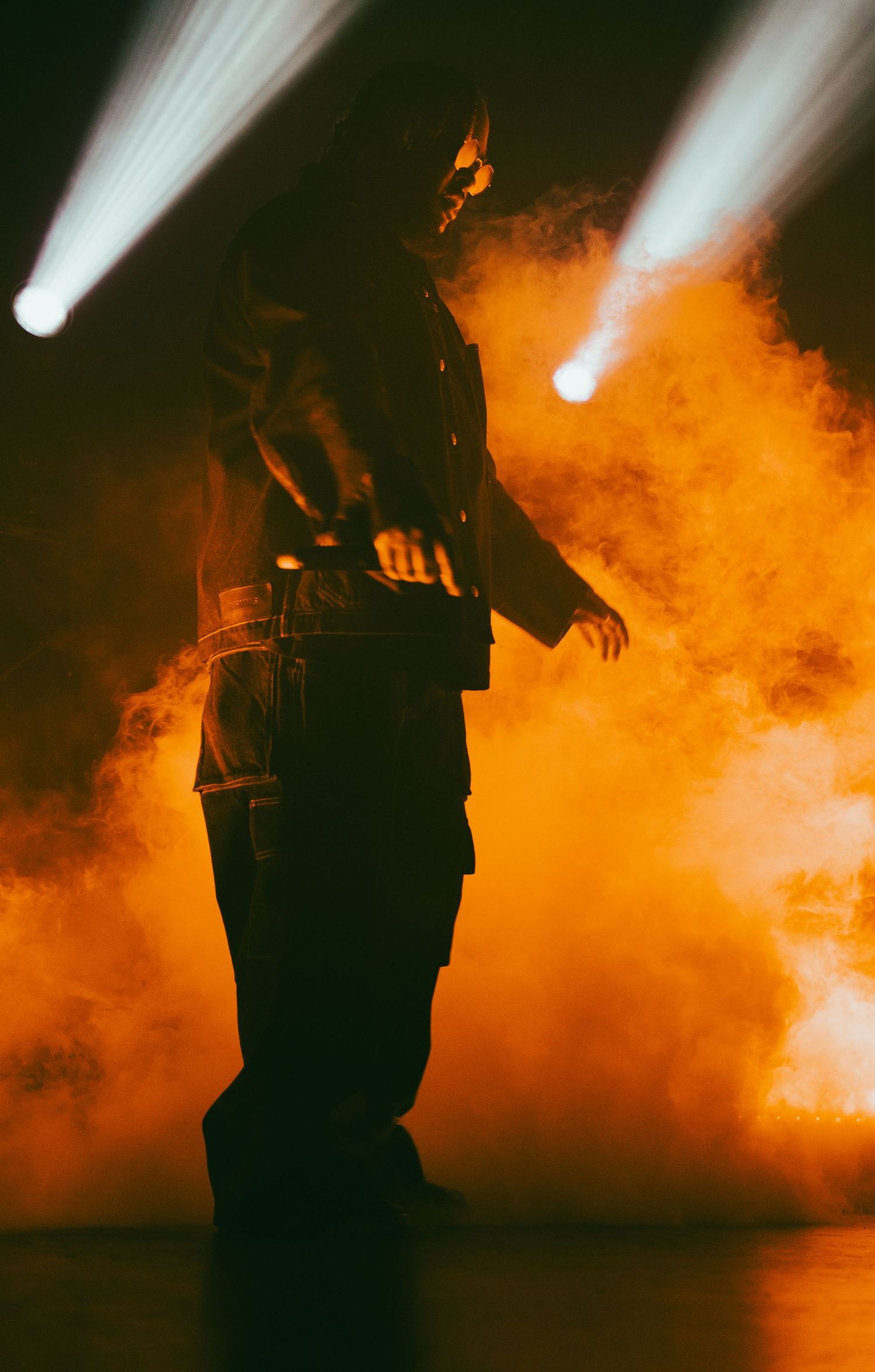 Silhouetted performer standing in dramatic orange smoke with bright stage spotlights overhead.