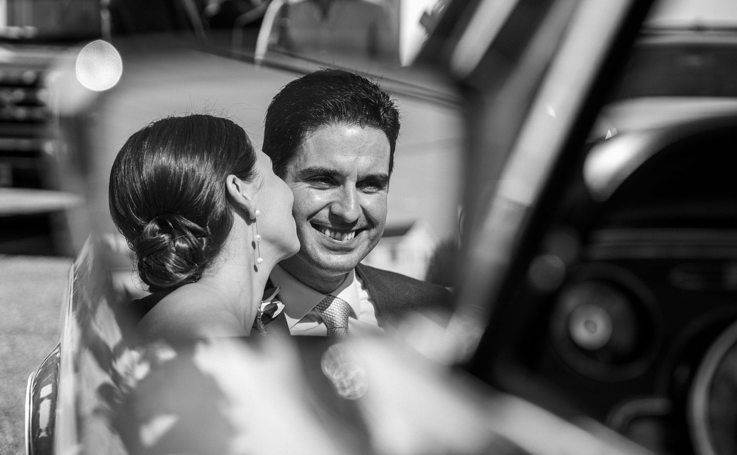 Groom smiling warmly at bride reflected in vintage car window in artistic black and white wedding photo.