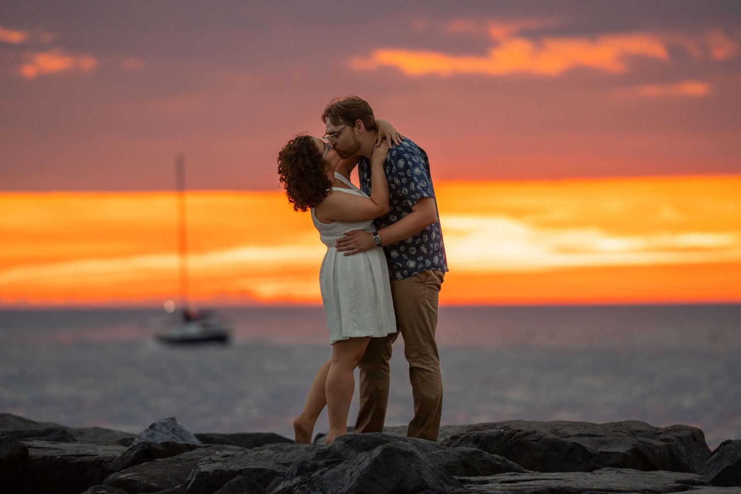 Couple kissing on rocky shoreline at sunset with sailboat and vibrant orange sky in background.