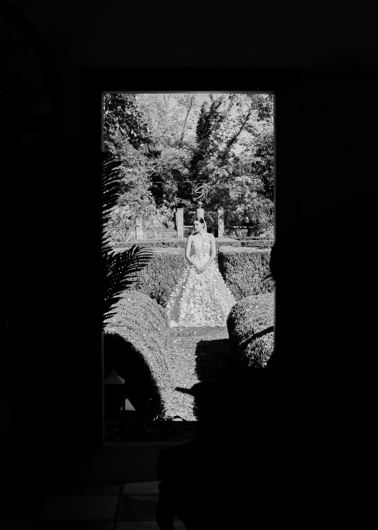A black and white framed view of a bride in a floral gown standing in a lush garden pathway.