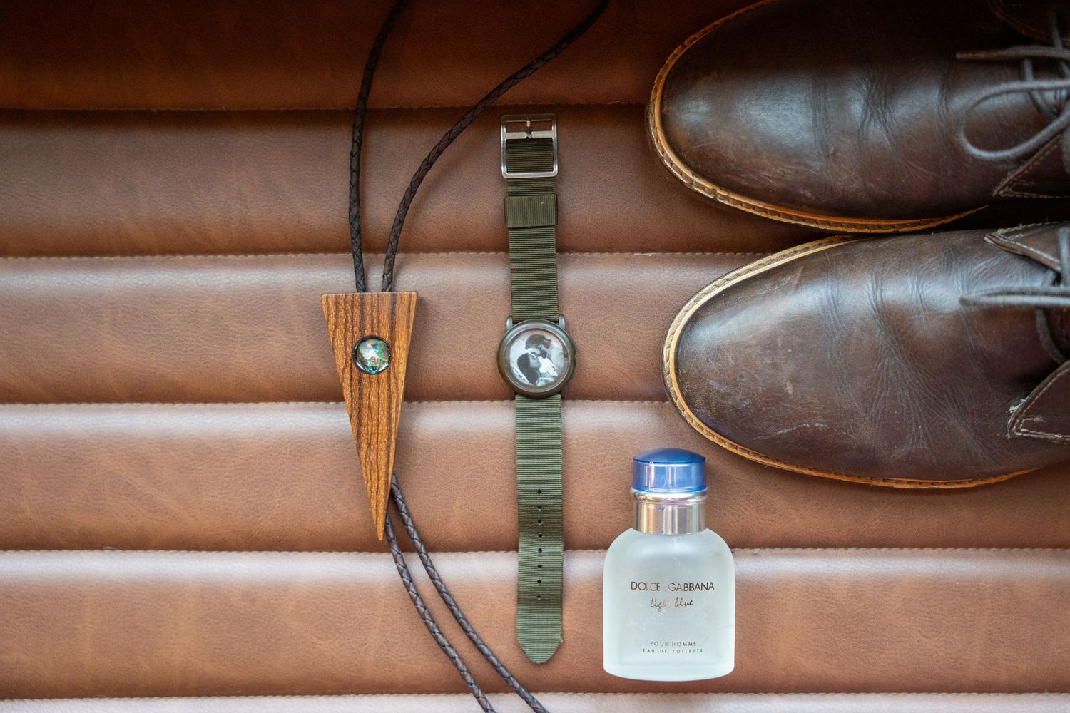 Groom wedding details flat lay featuring brown leather shoes, green NATO watch, bolo tie, and cologne.