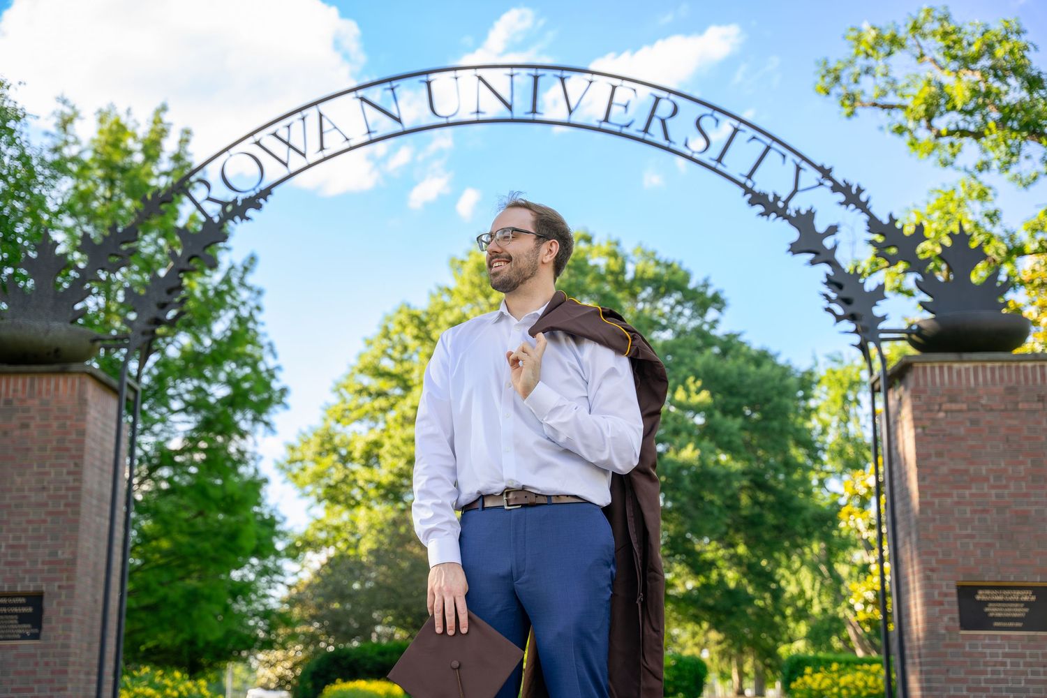 Male graduate in dress shirt holding gown over shoulder at Rowan University gate.