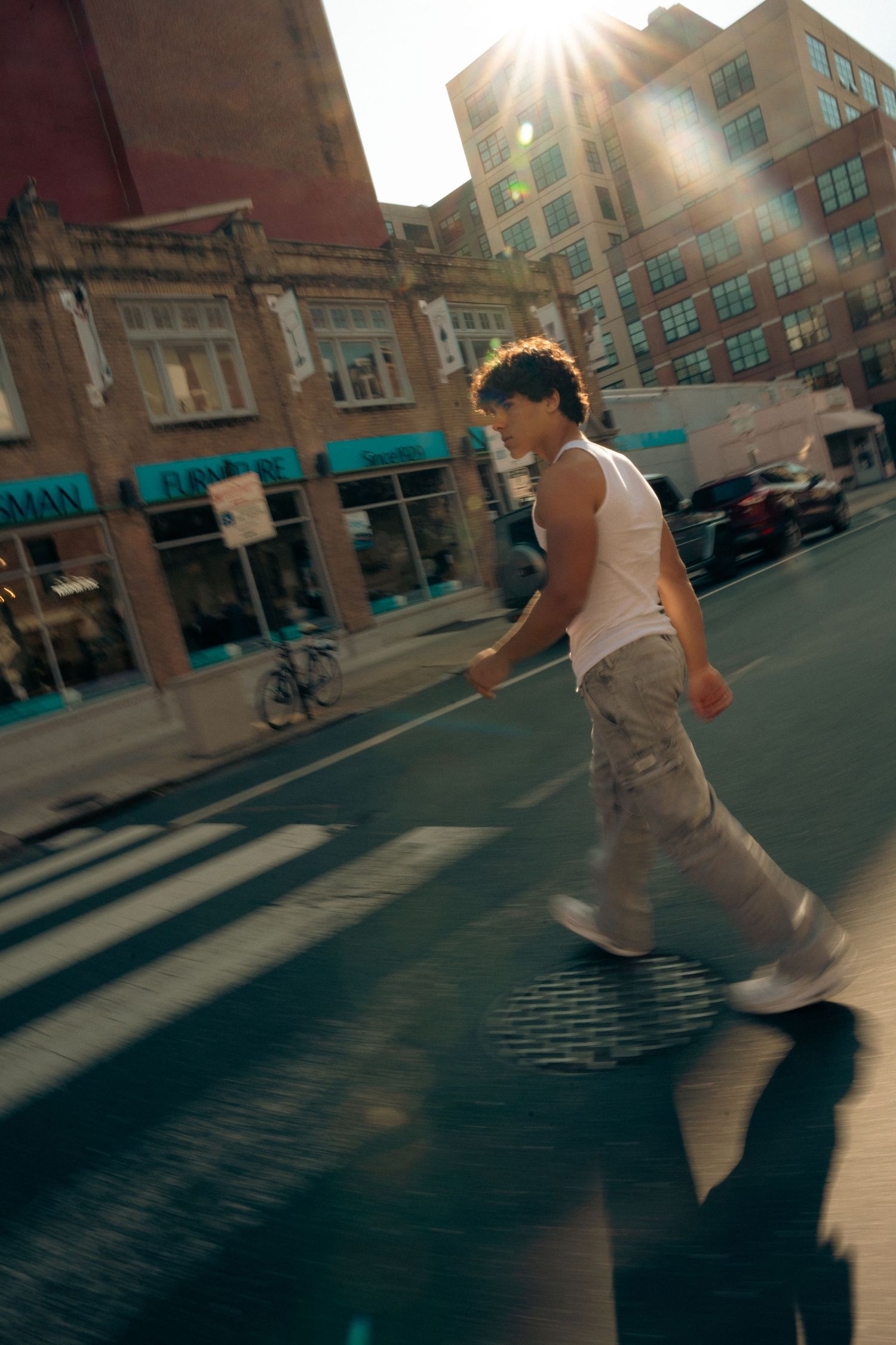 Person in white tank top crossing sunny urban street with lens flare and motion blur.