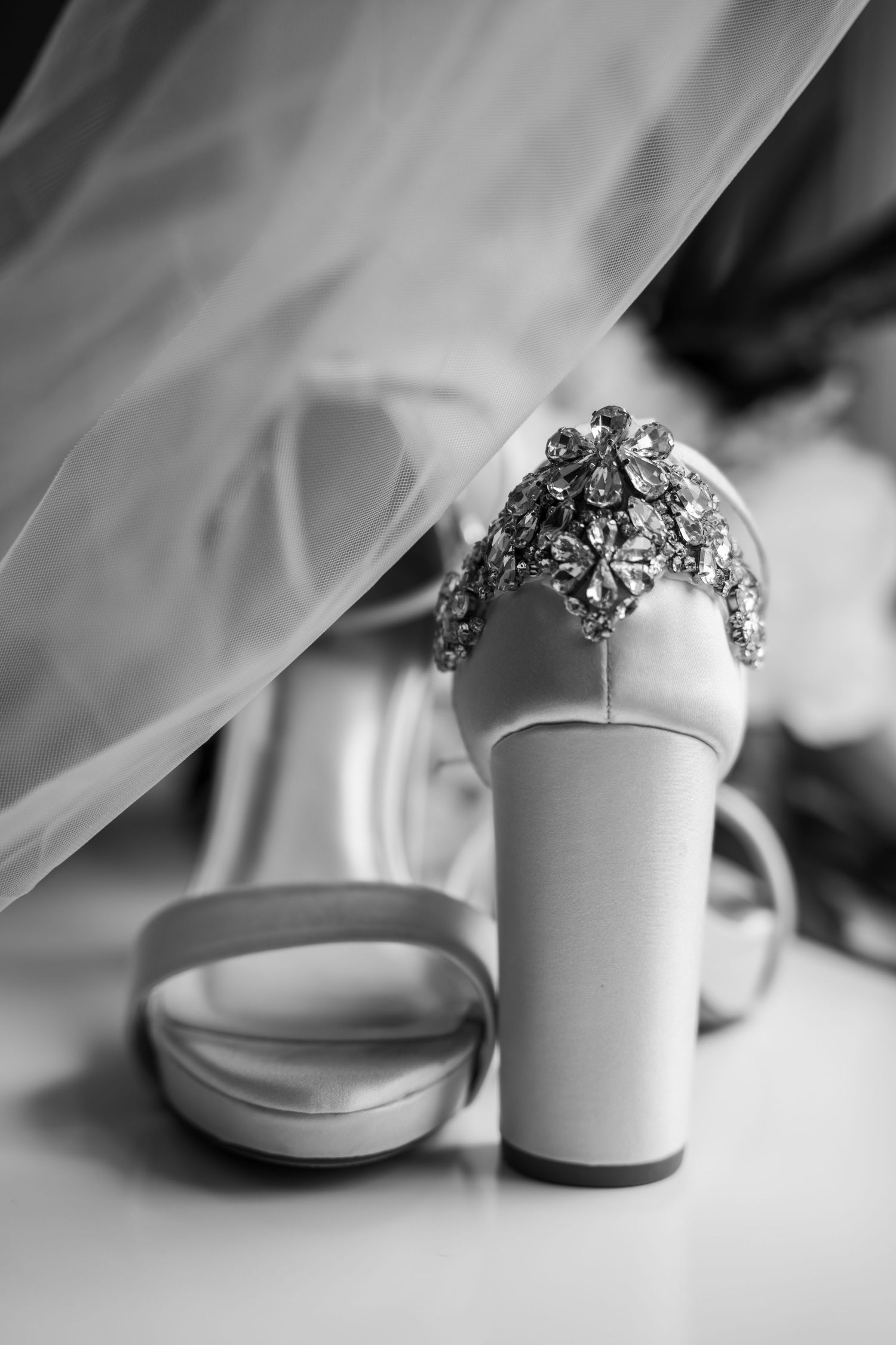 Black and white close-up of jeweled bridal heels with crystal embellishments beneath a wedding veil.