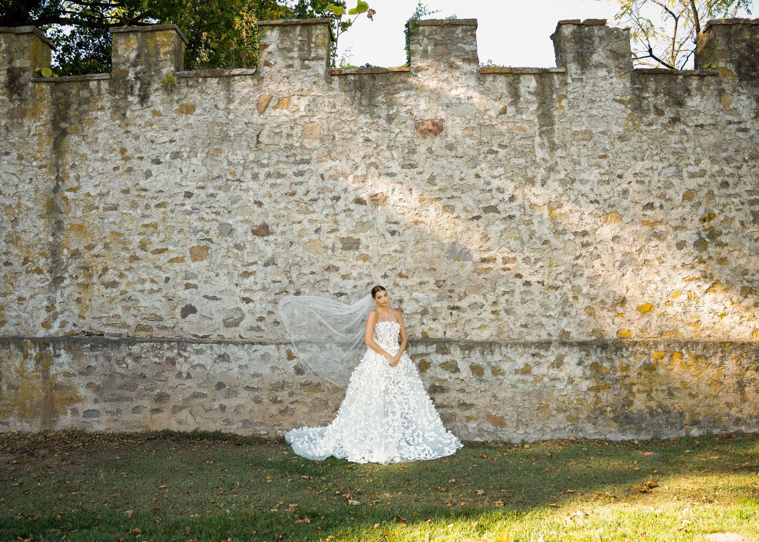 Elegant bride in lace ball gown poses gracefully before ancient stone wall in outdoor setting.