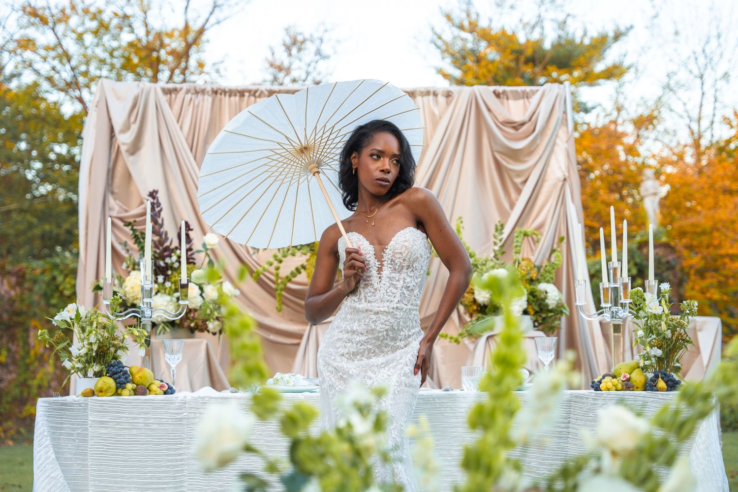 Elegant bride in fitted lace gown holds parasol beside decorated sweetheart table at outdoor wedding.
