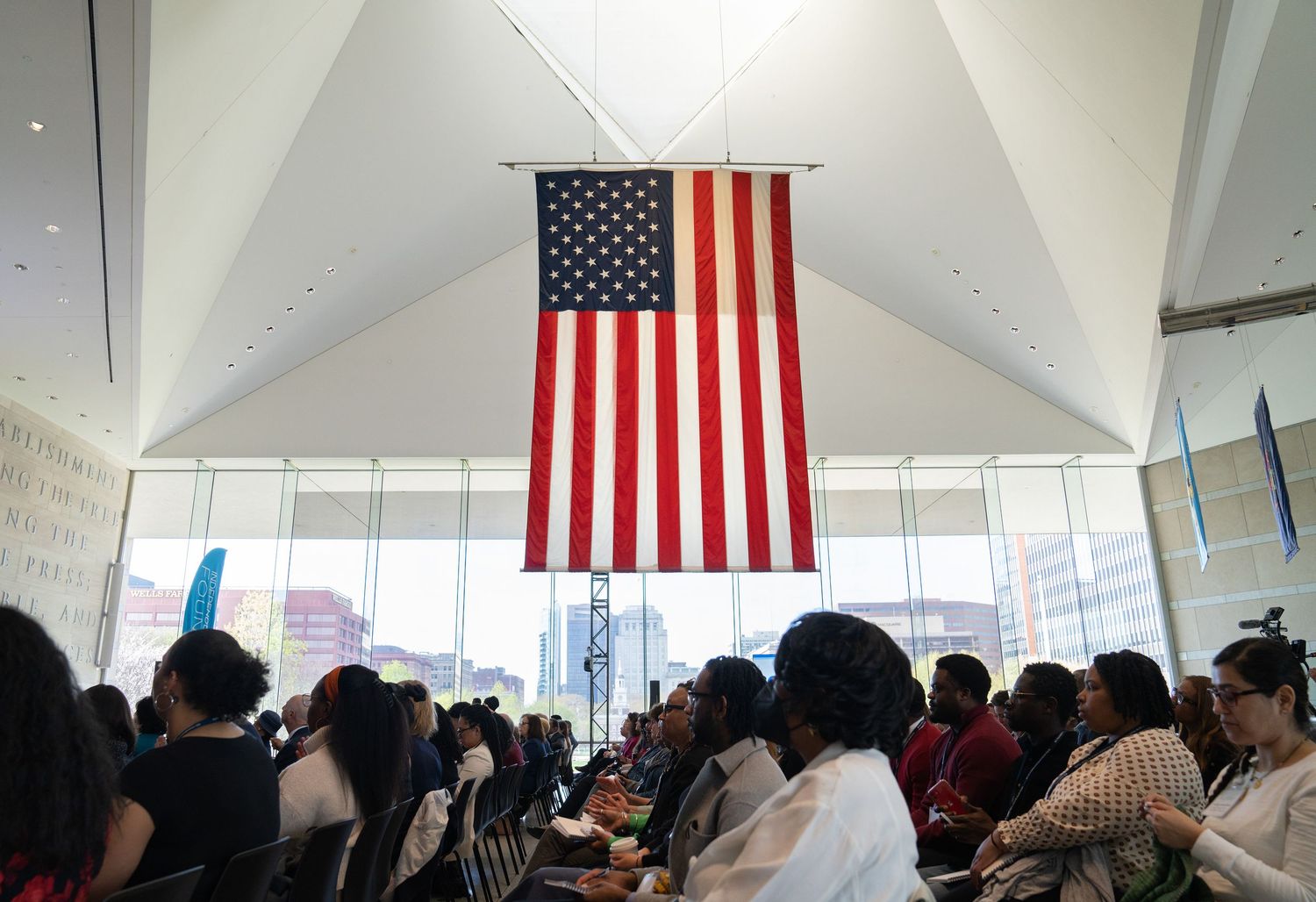 Large American flag hanging in modern building atrium above diverse seated crowd.