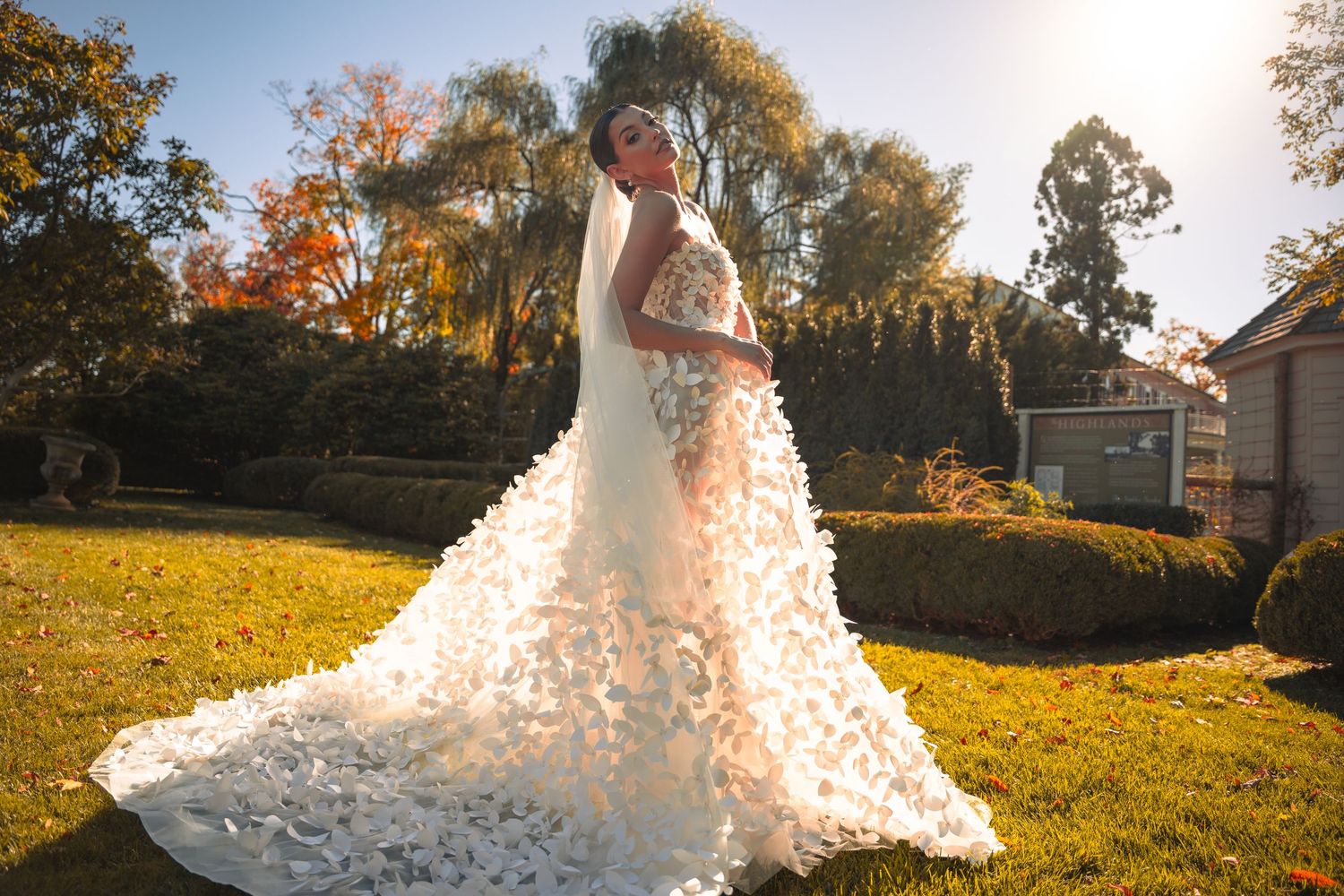 Bride in stunning white floral applique wedding gown with long train poses outdoors in sunlit garden.