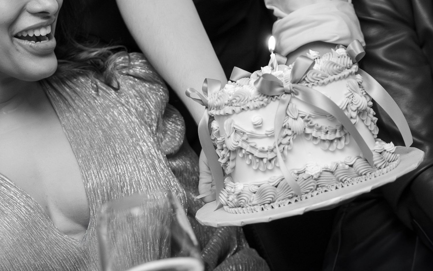 Black and white photo of a decorated celebration cake with a candle being held at a party.